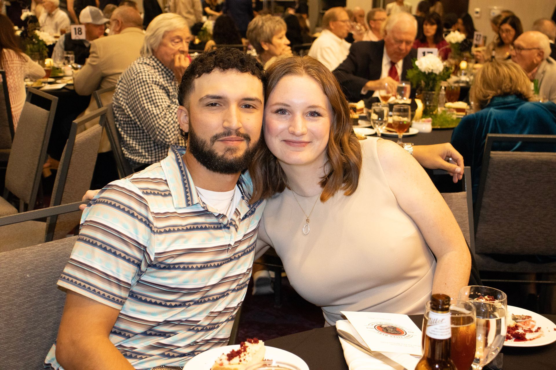 A couple smiles together while sitting at a table in a banquet hall, with other guests visible in the blurred background.