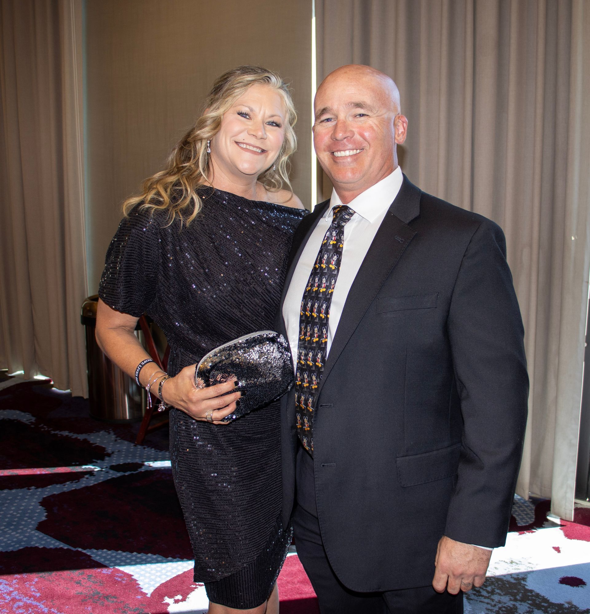 A woman in a black sequined dress and a man in a suit and patterned tie smile while standing together indoors.