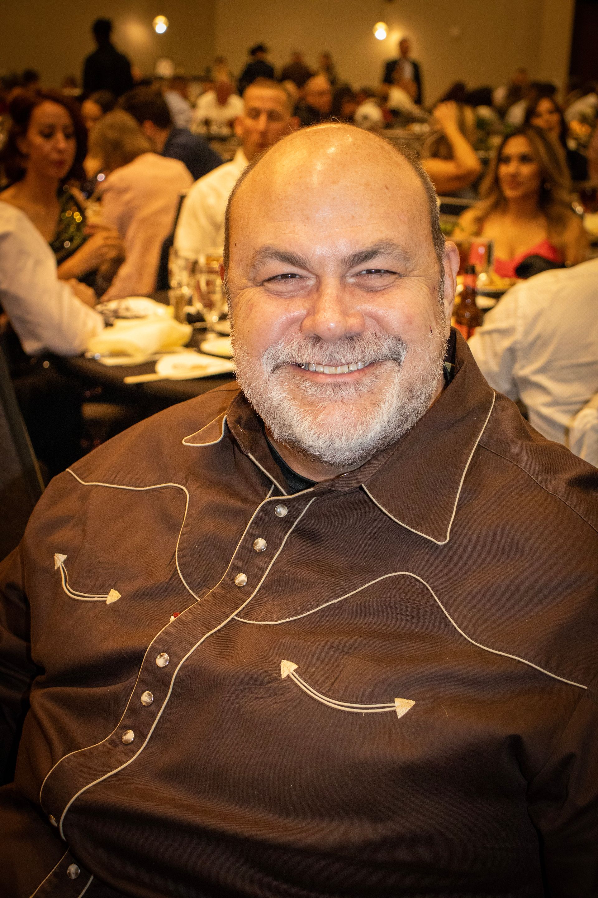 Smiling person in a dark brown western-style button-down shirt at a crowded event.