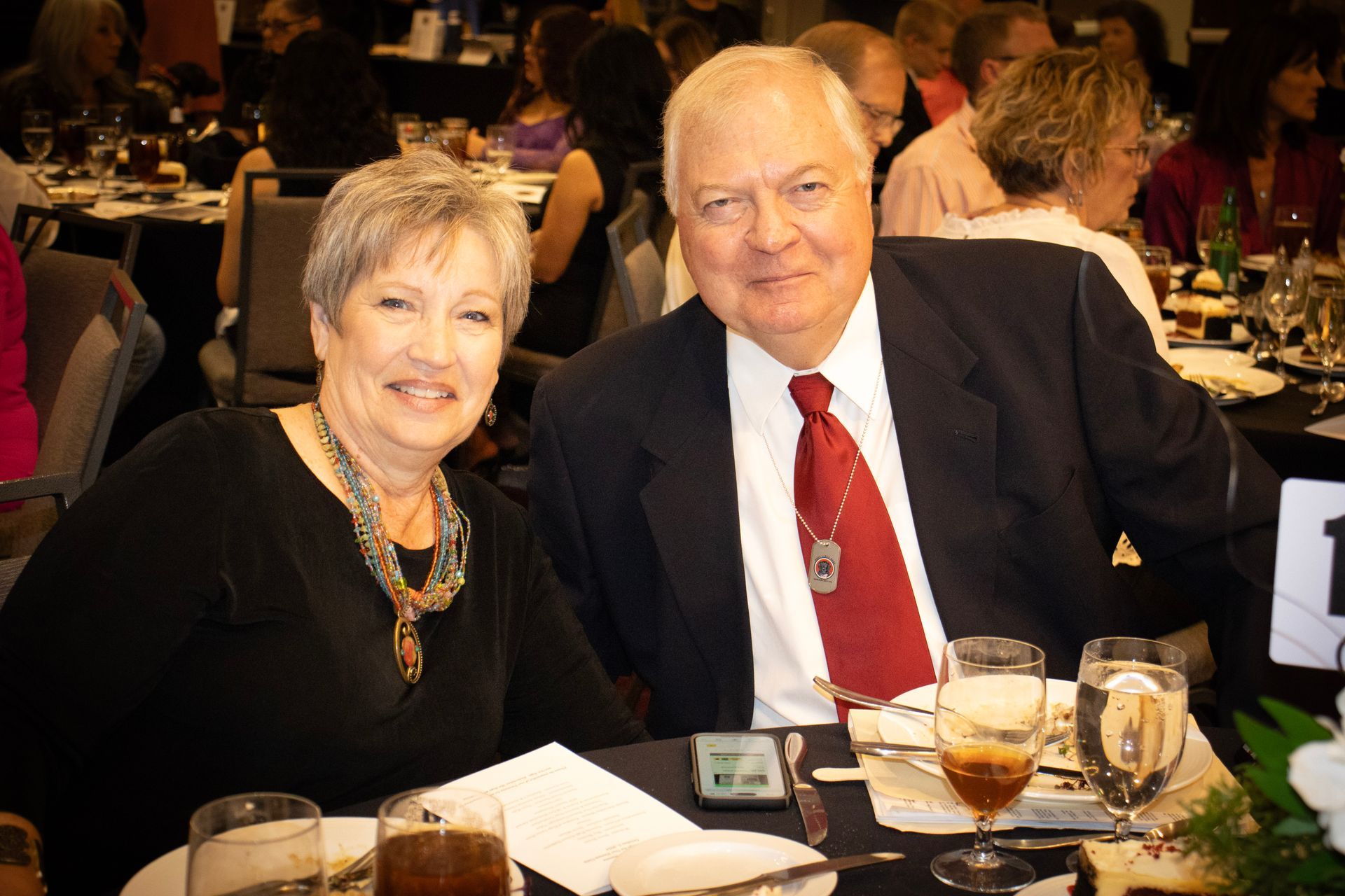 An older couple posing at a table during a dinner event, smiling towards the camera with glasses and place settings nearby.
