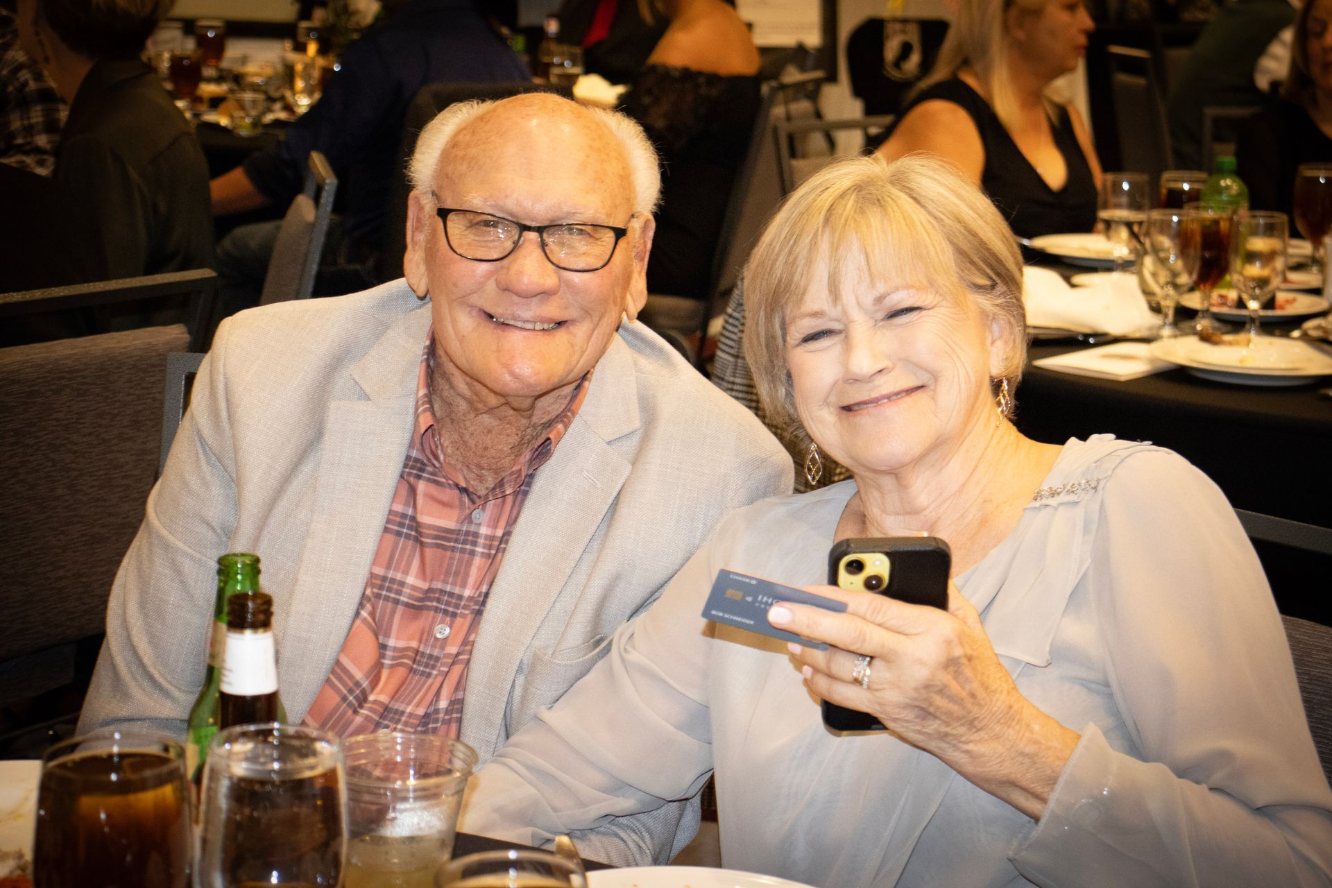 An older couple smiles at the camera at a table in a restaurant; the woman holds a credit card and smartphone.