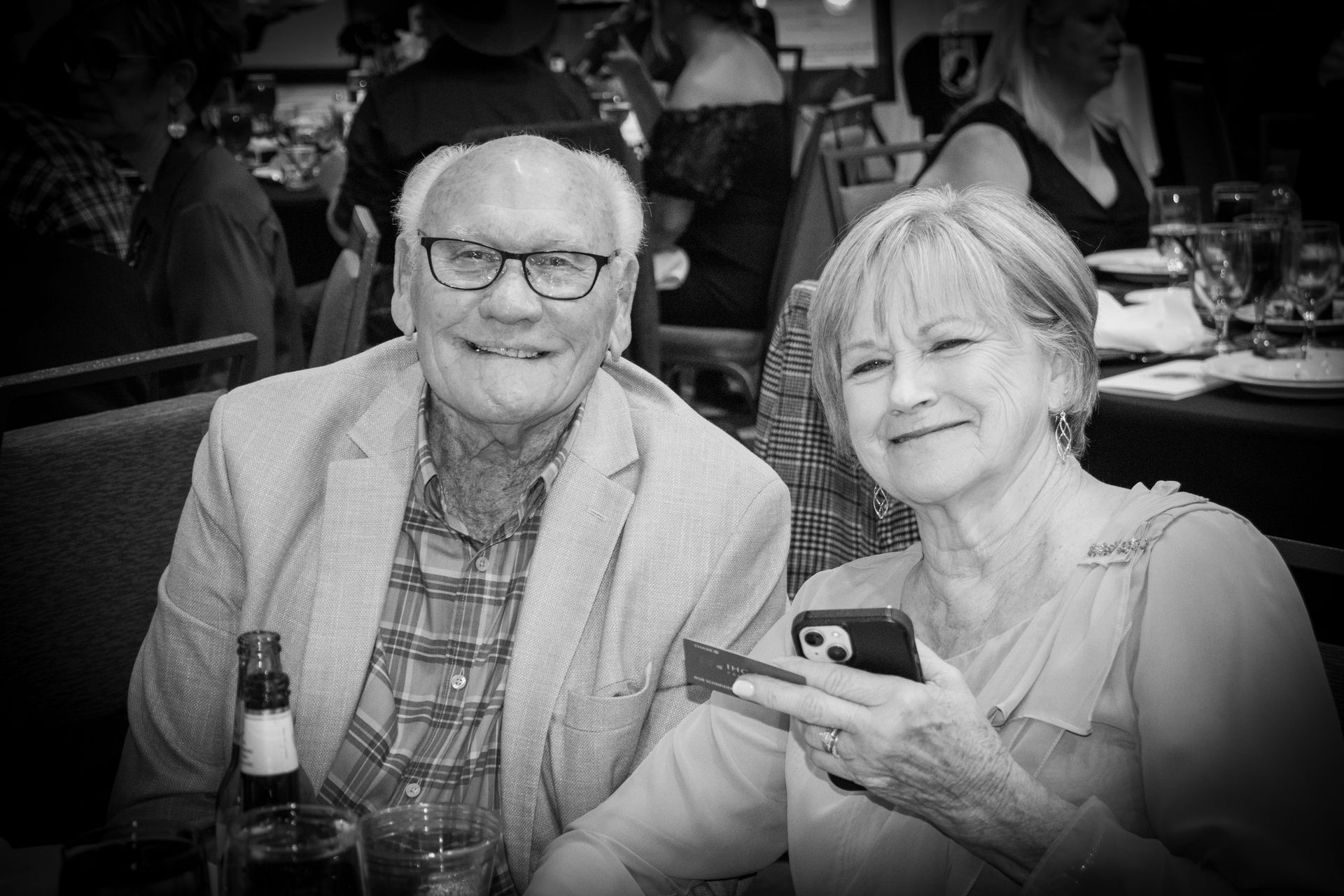 An older couple smiles for a photo at an indoor dining event; the person on the right holds a smartphone.