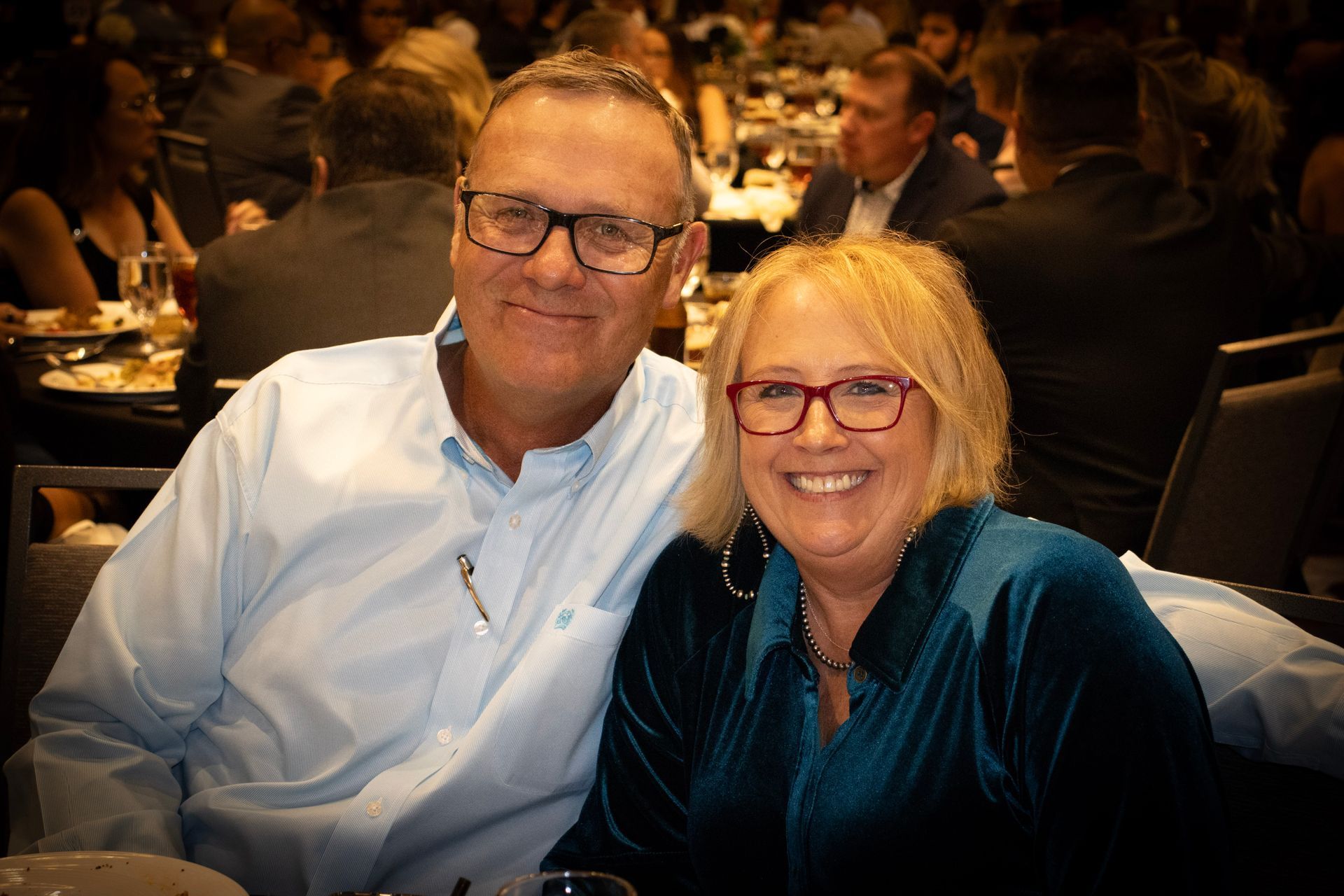 A smiling couple with glasses sits close together at a dimly lit indoor event with other guests in the background.