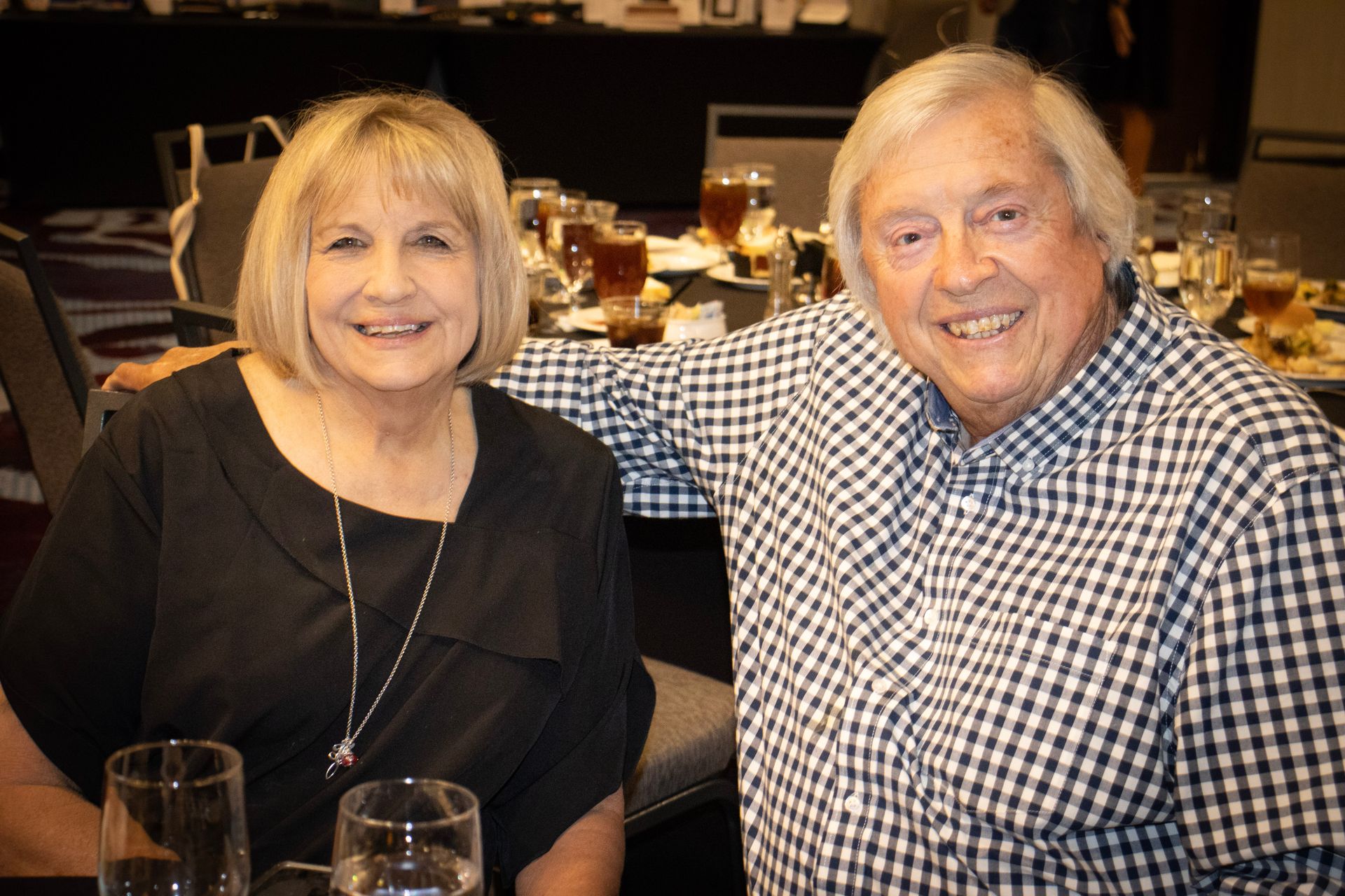 A smiling couple sitting side-by-side at a table in a dimly lit, event-style setting.