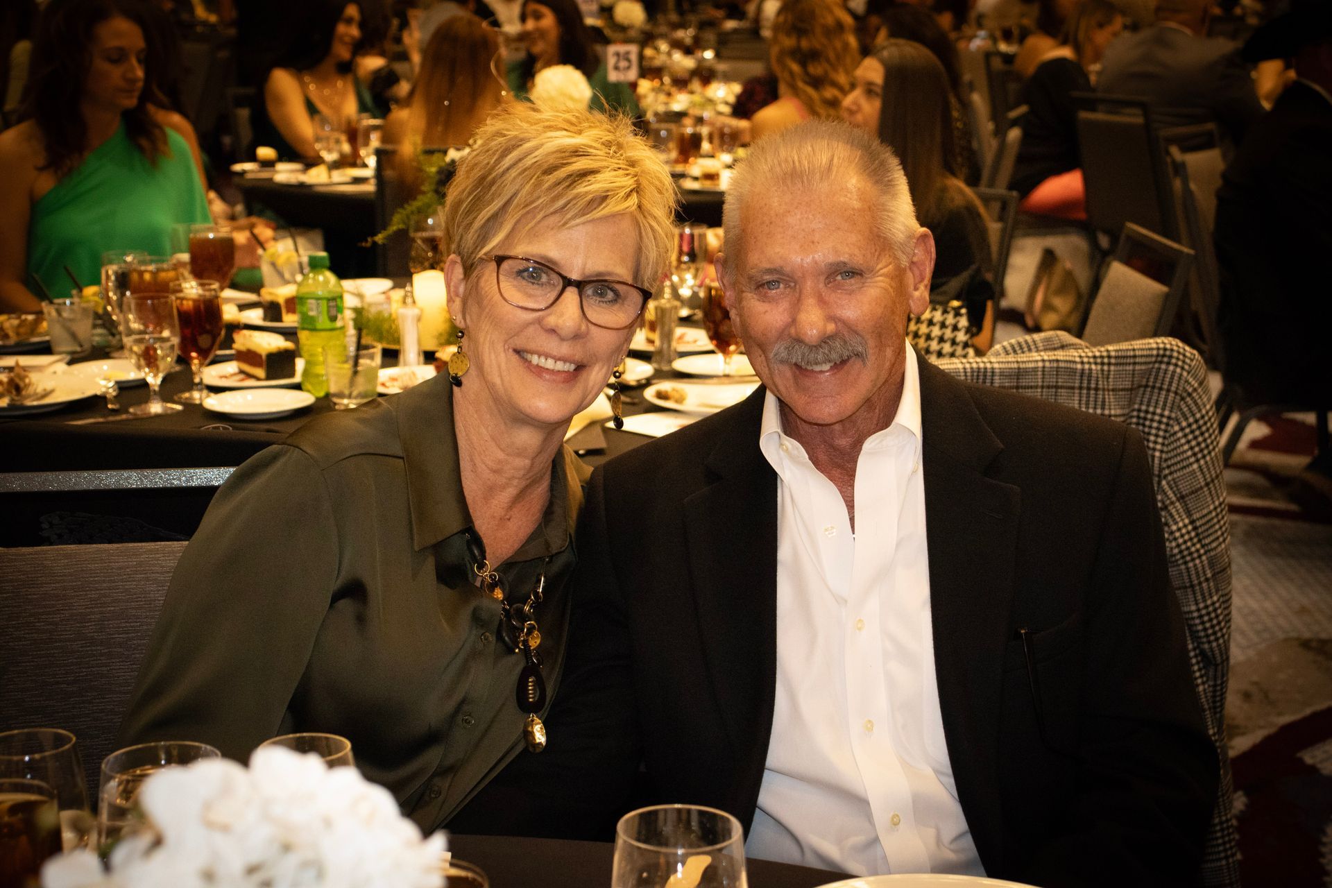 A smiling couple poses for a photo at a dimly lit banquet hall, sitting at a table with drinks and a floral centerpiece.