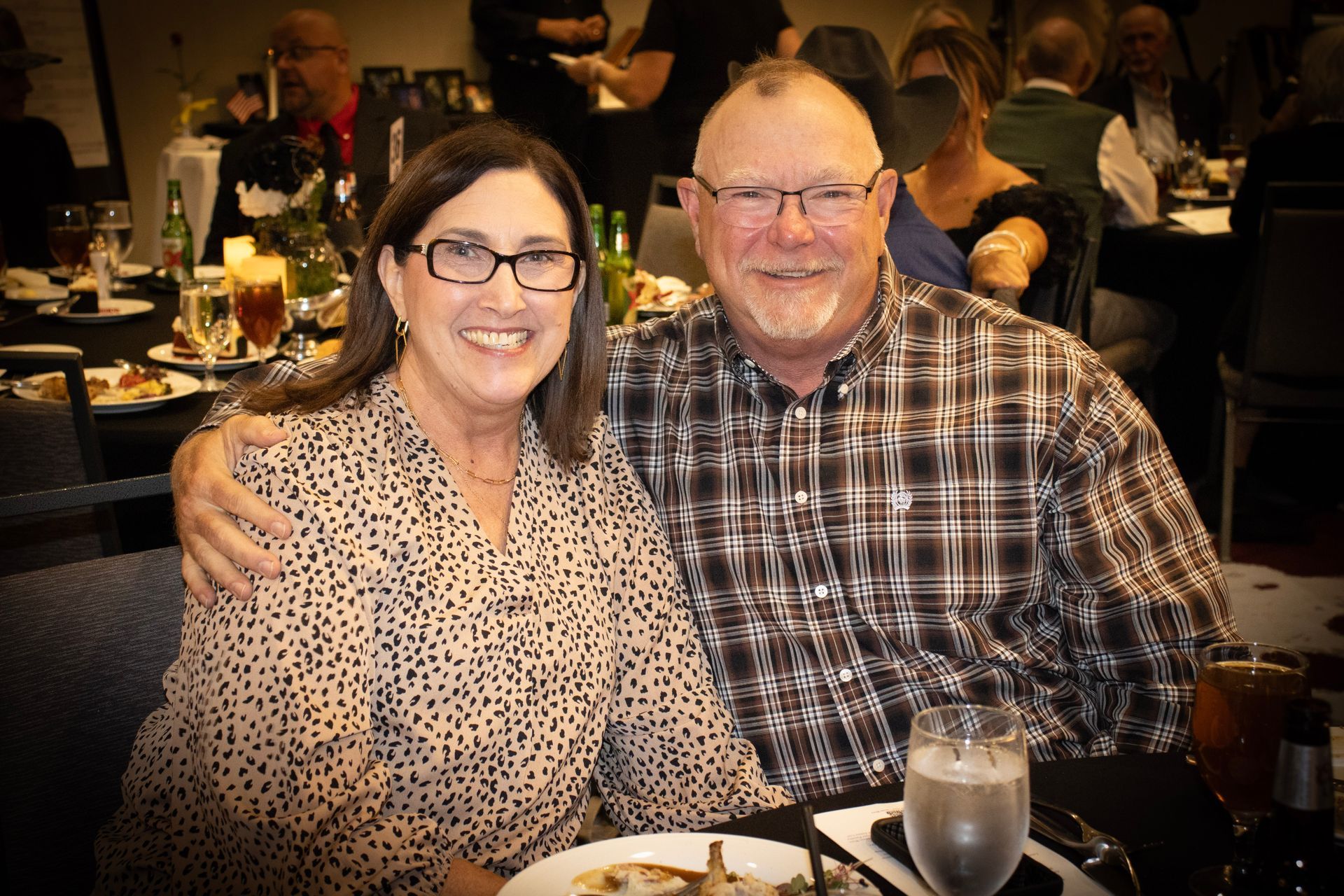 A smiling couple poses for a photo at a table in a dimly lit restaurant, the man with his arm around the woman's shoulder.