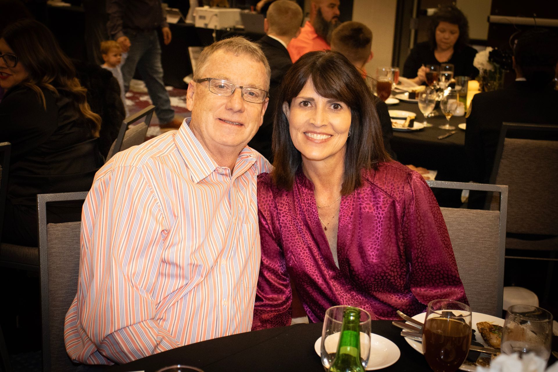 A couple smiles while sitting at a table with drinks and plates in a dimly lit event space.