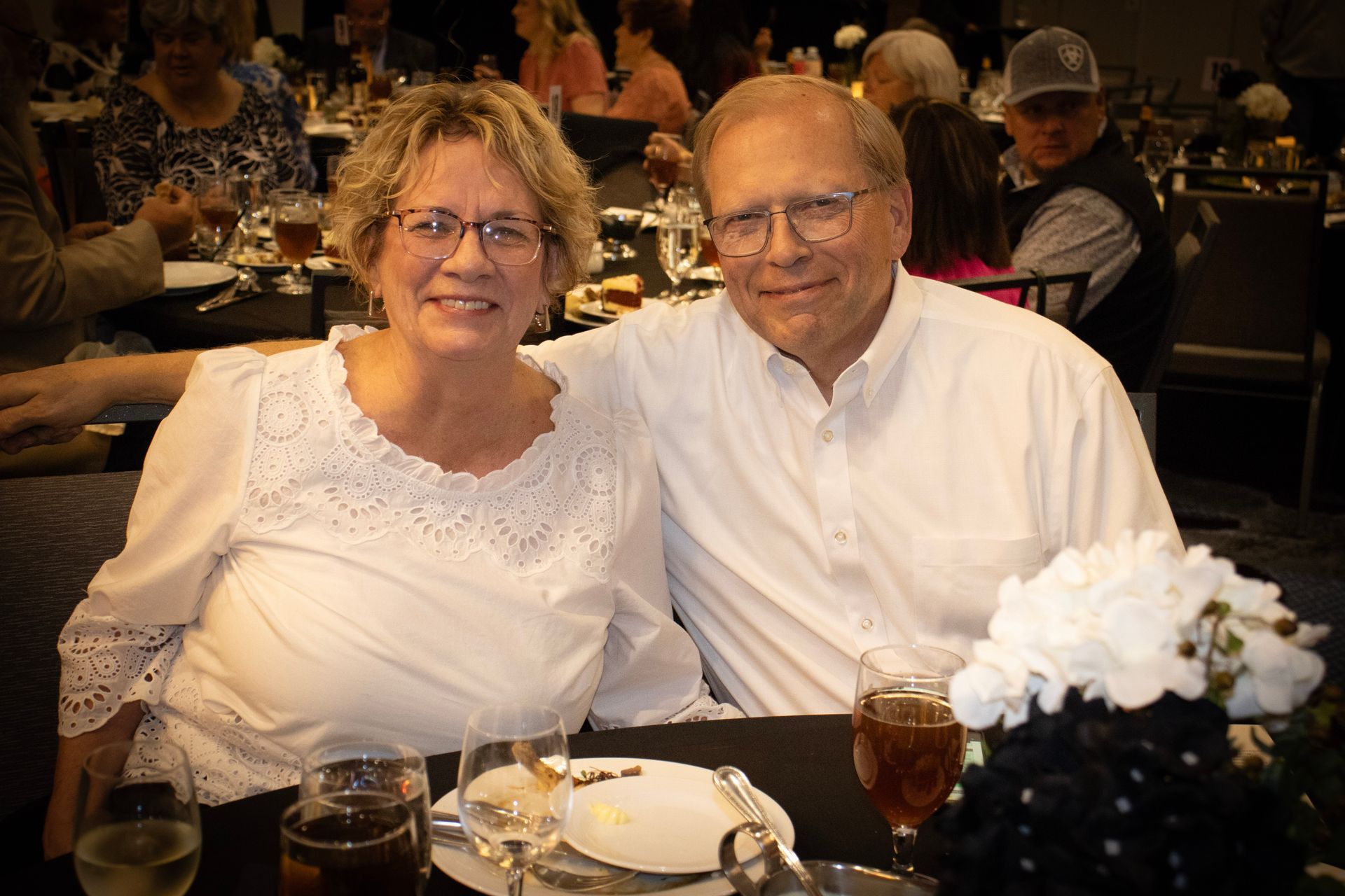 A smiling couple sits at a dining table at an event, with other guests visible in the background.
