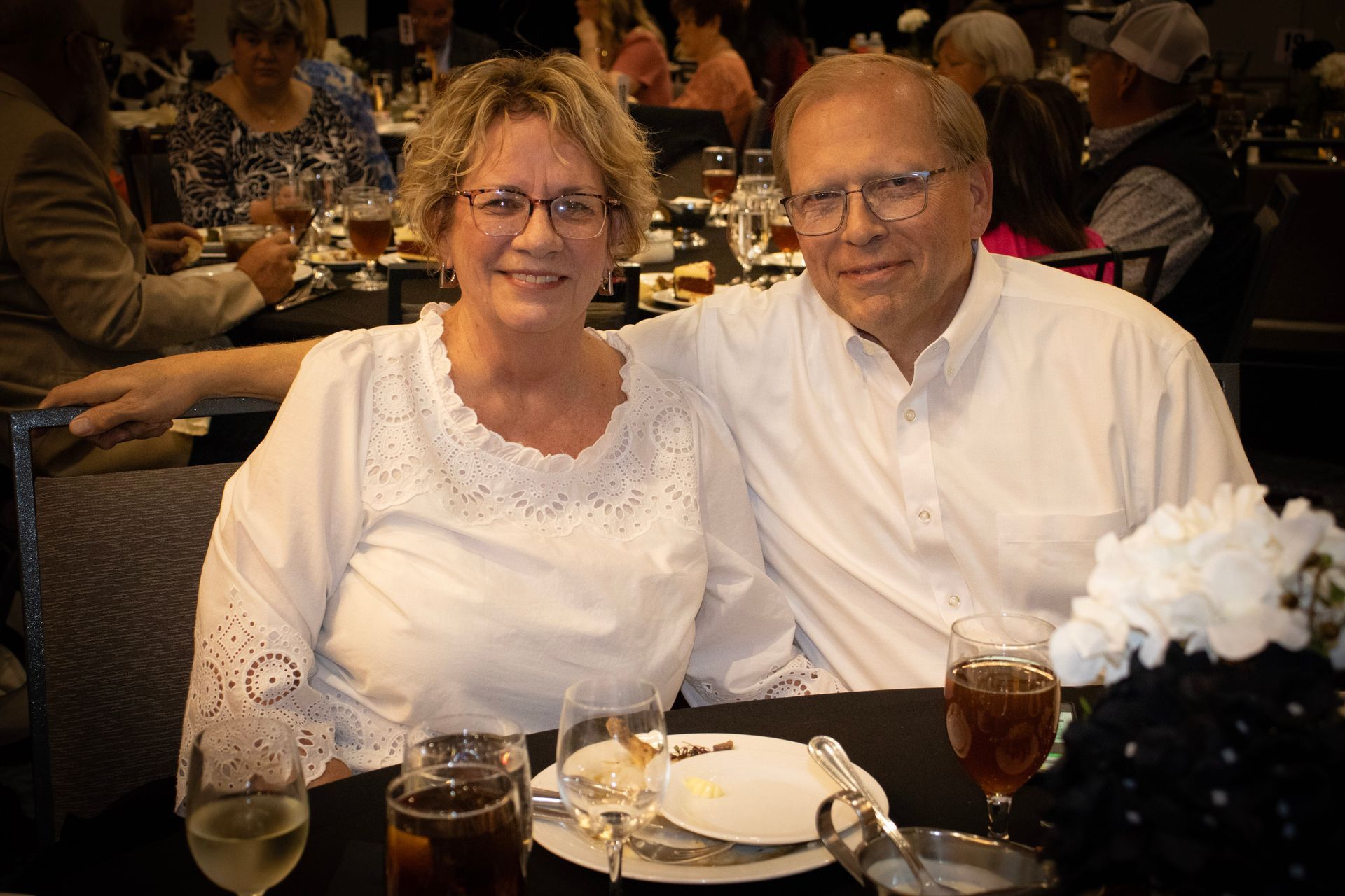 A couple in matching white tops sit together at a table in a dimly lit banquet hall, smiling for the camera.