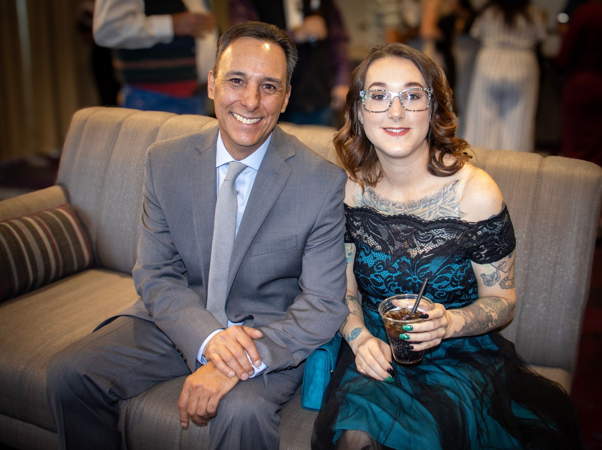 A man in a suit and a woman in a dark dress with tattoos sit on a grey sofa at an indoor event, smiling at the camera.