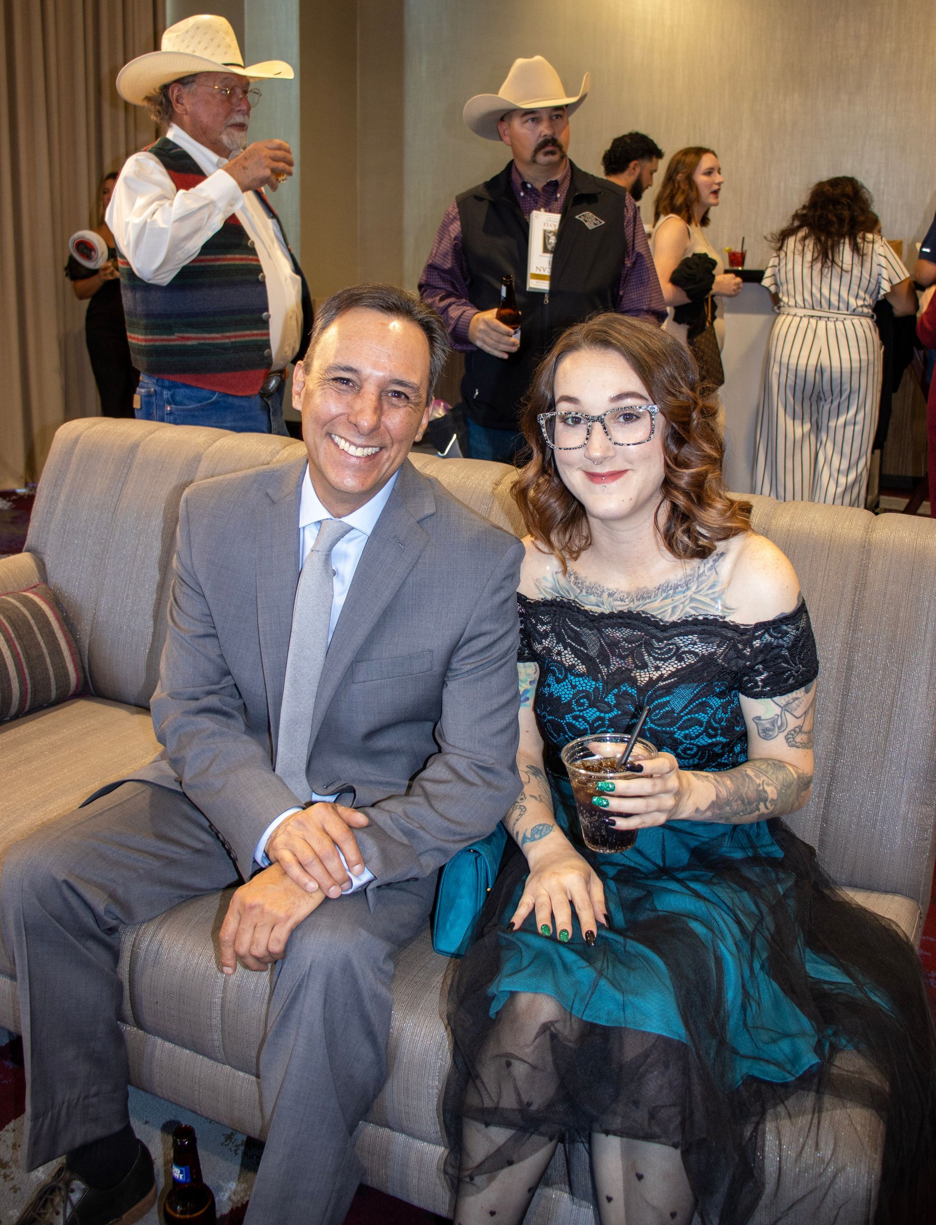 A smiling man in a gray suit and a woman in a dark teal dress sit together on a sofa, with others in the background.