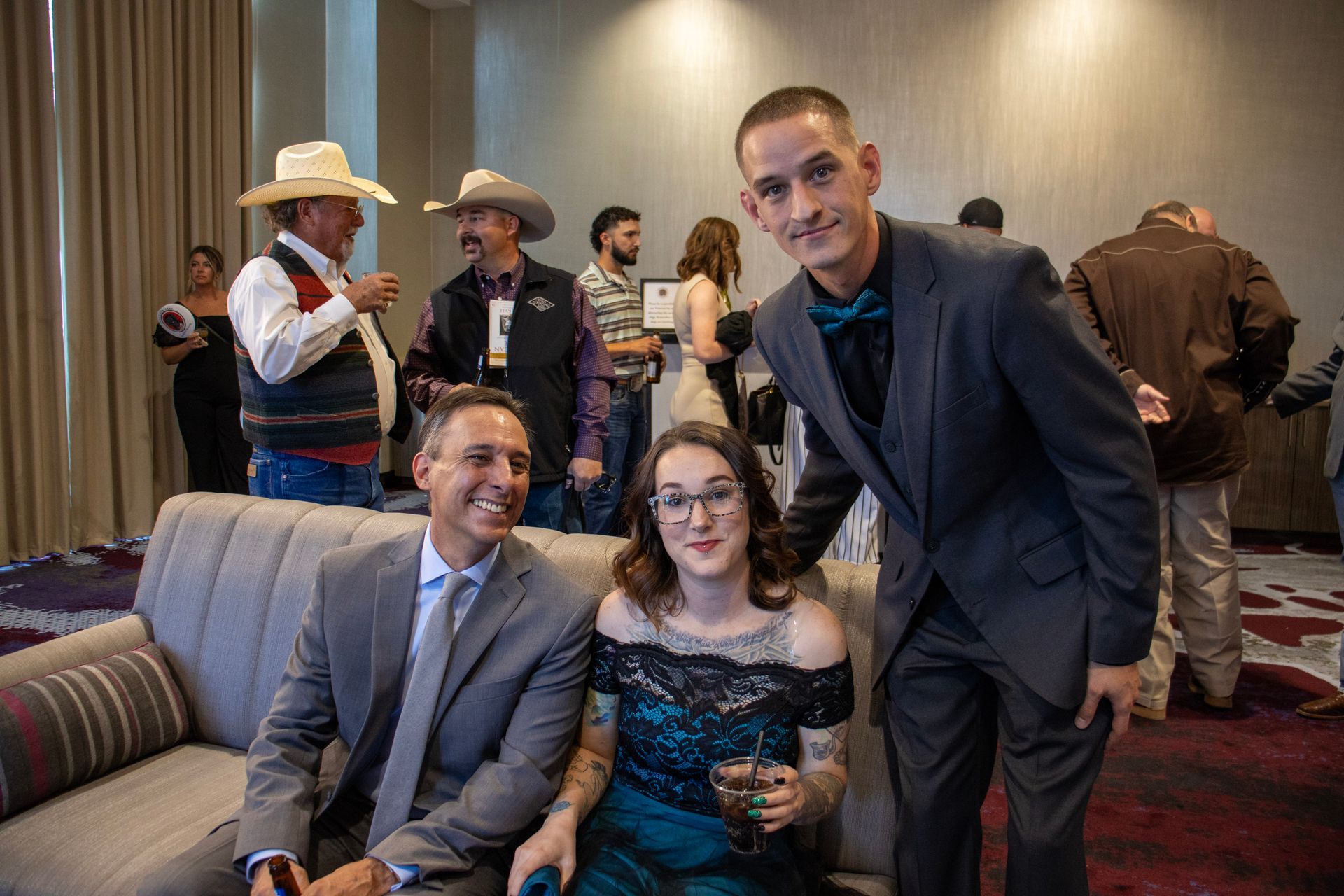 A group of people in formal and semi-formal attire gather in a room, with three people smiling on a sofa and standing.