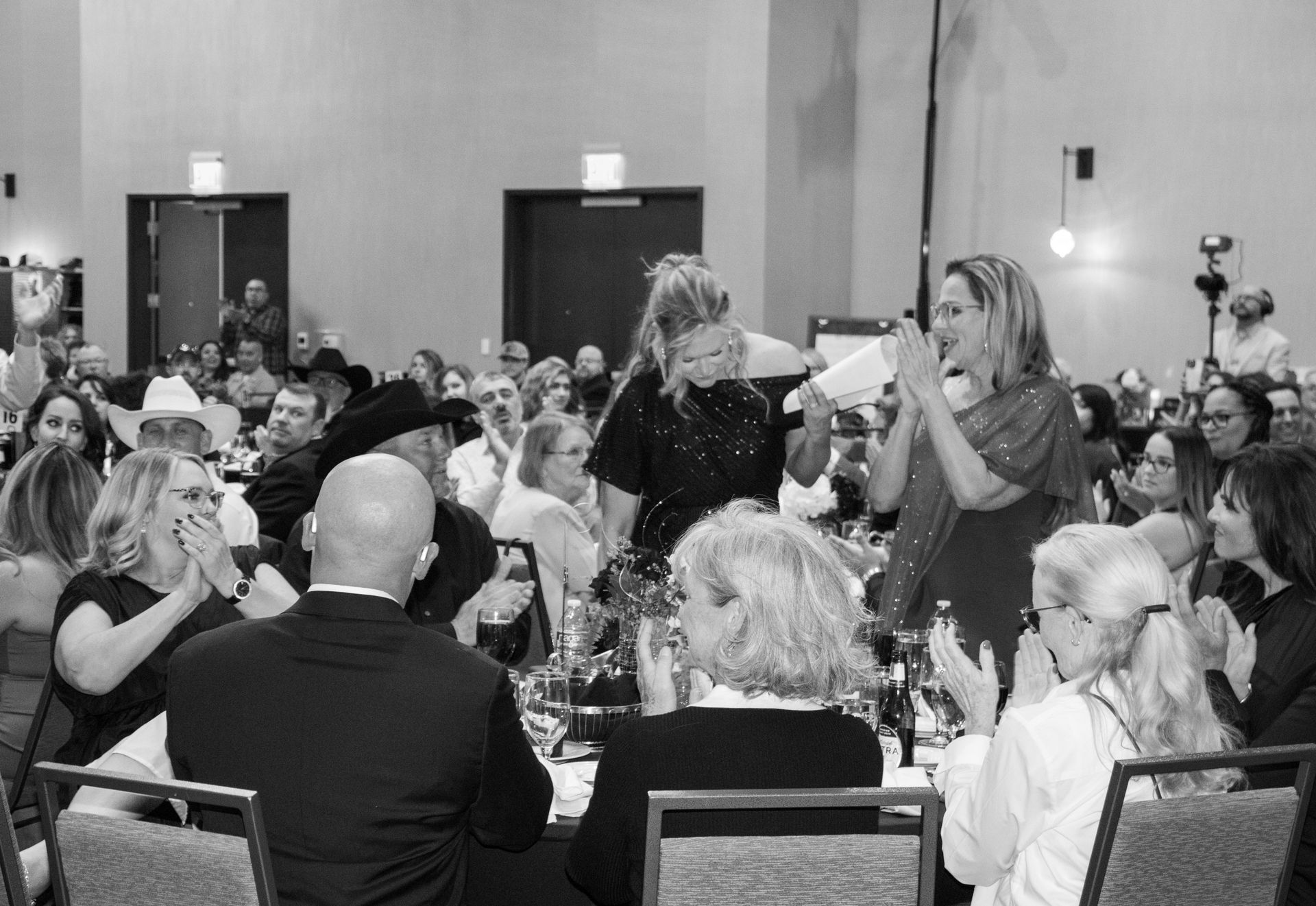 Attendees applaud and react at a formal indoor event, with a woman standing and speaking near the center of the room.