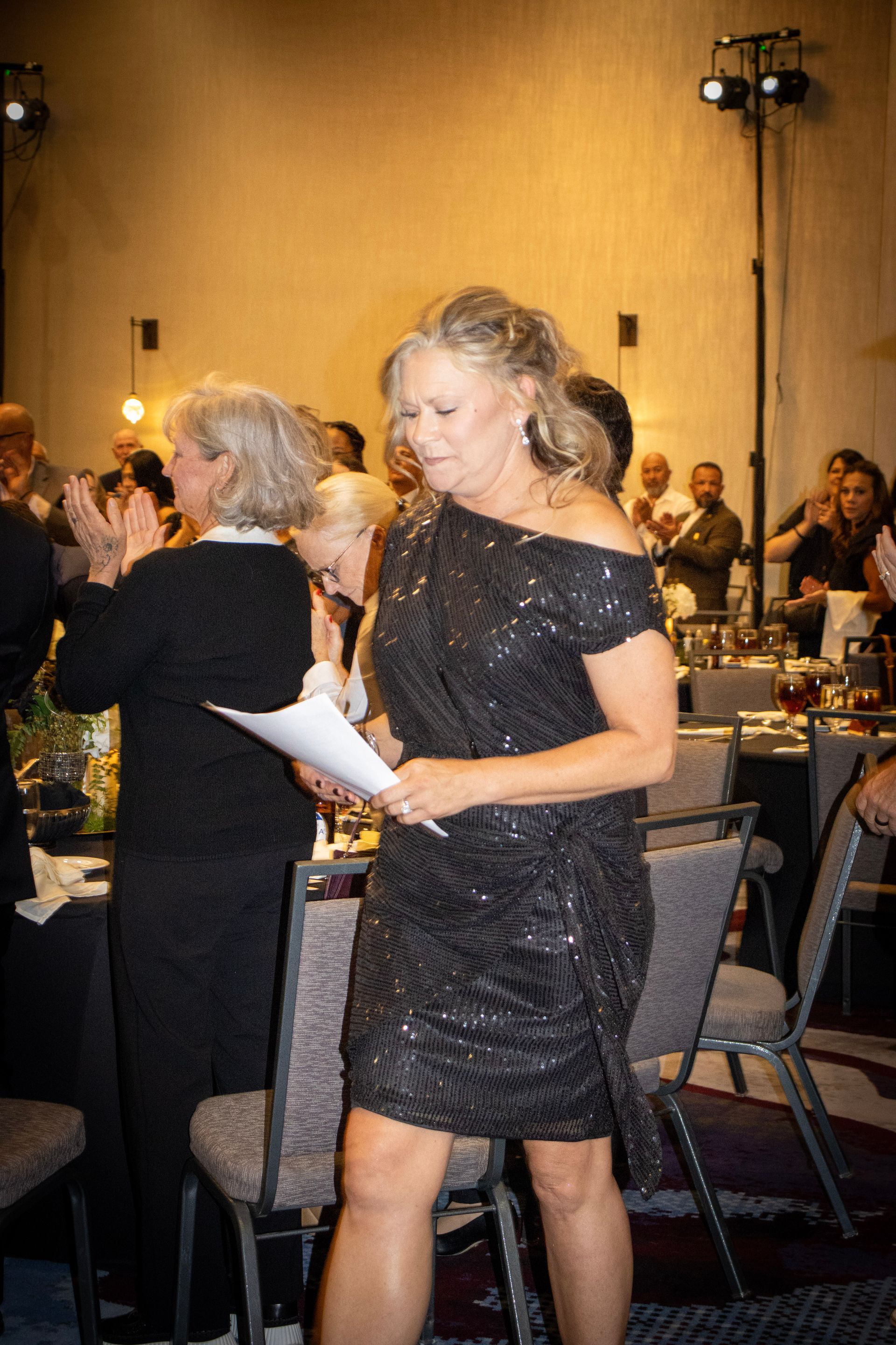 A person in a black sequined dress holds papers while walking through a room with tables and guests in the background.