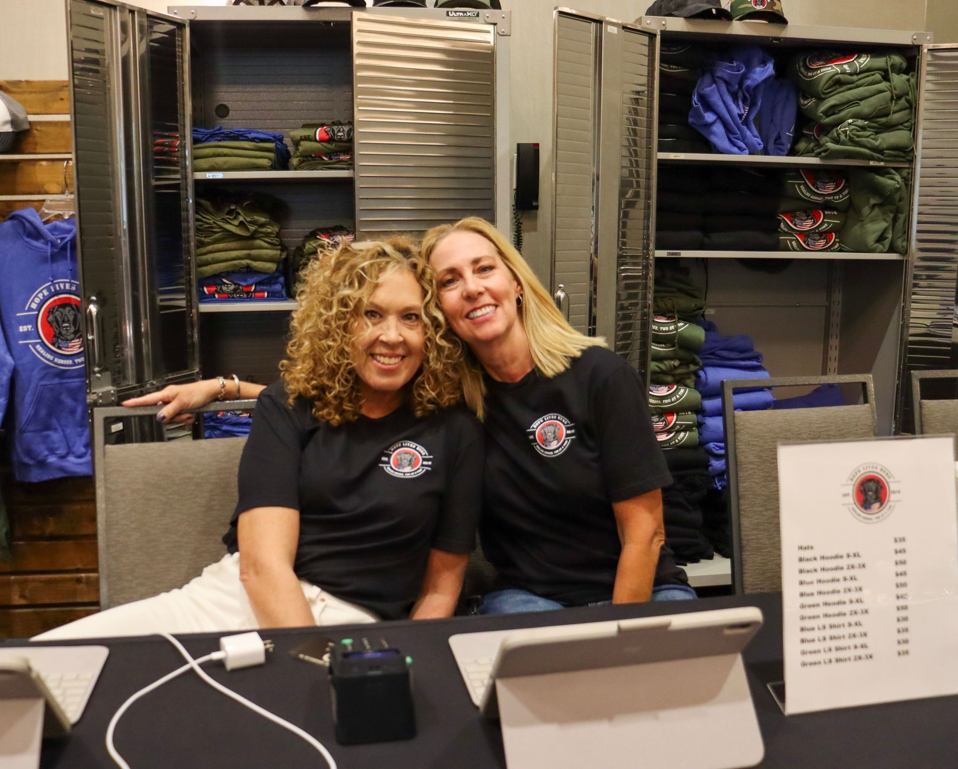 Two smiling people wearing matching black t-shirts sit behind a table with merchandise and a price list sign.