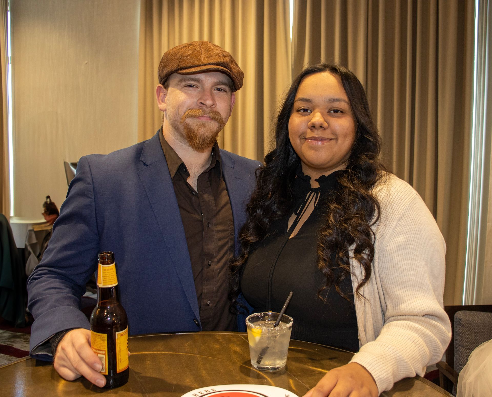 A man in a blue suit and cap, and a person in a black top and white cardigan stand at a table with drinks, smiling.