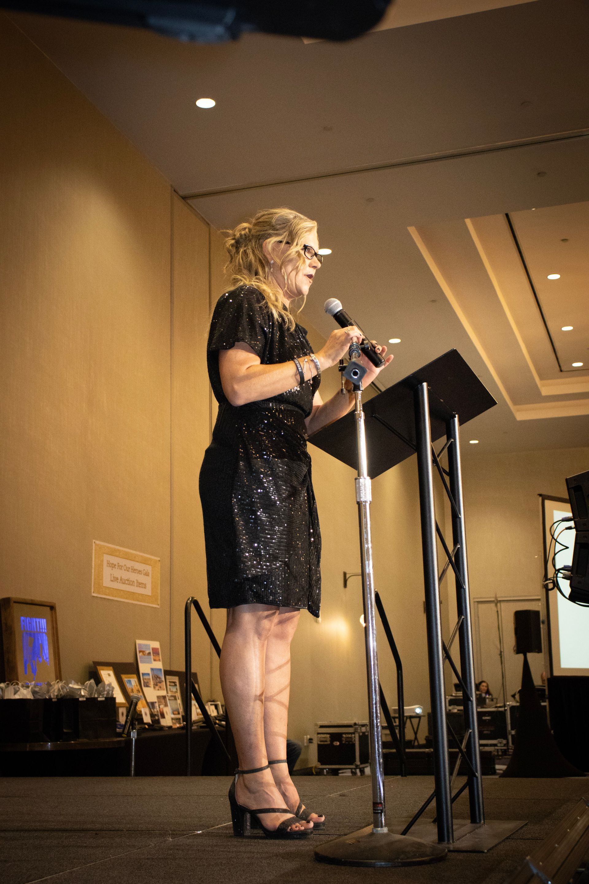 A woman in a black sequined dress stands behind a podium speaking into a microphone in an indoor event space.