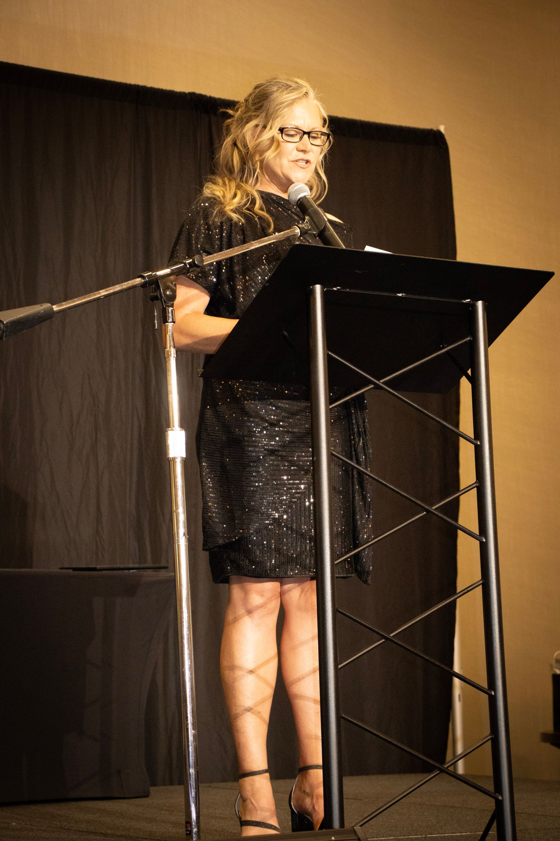A person in a black sequined dress stands at a podium in front of a dark curtain, speaking into a microphone.