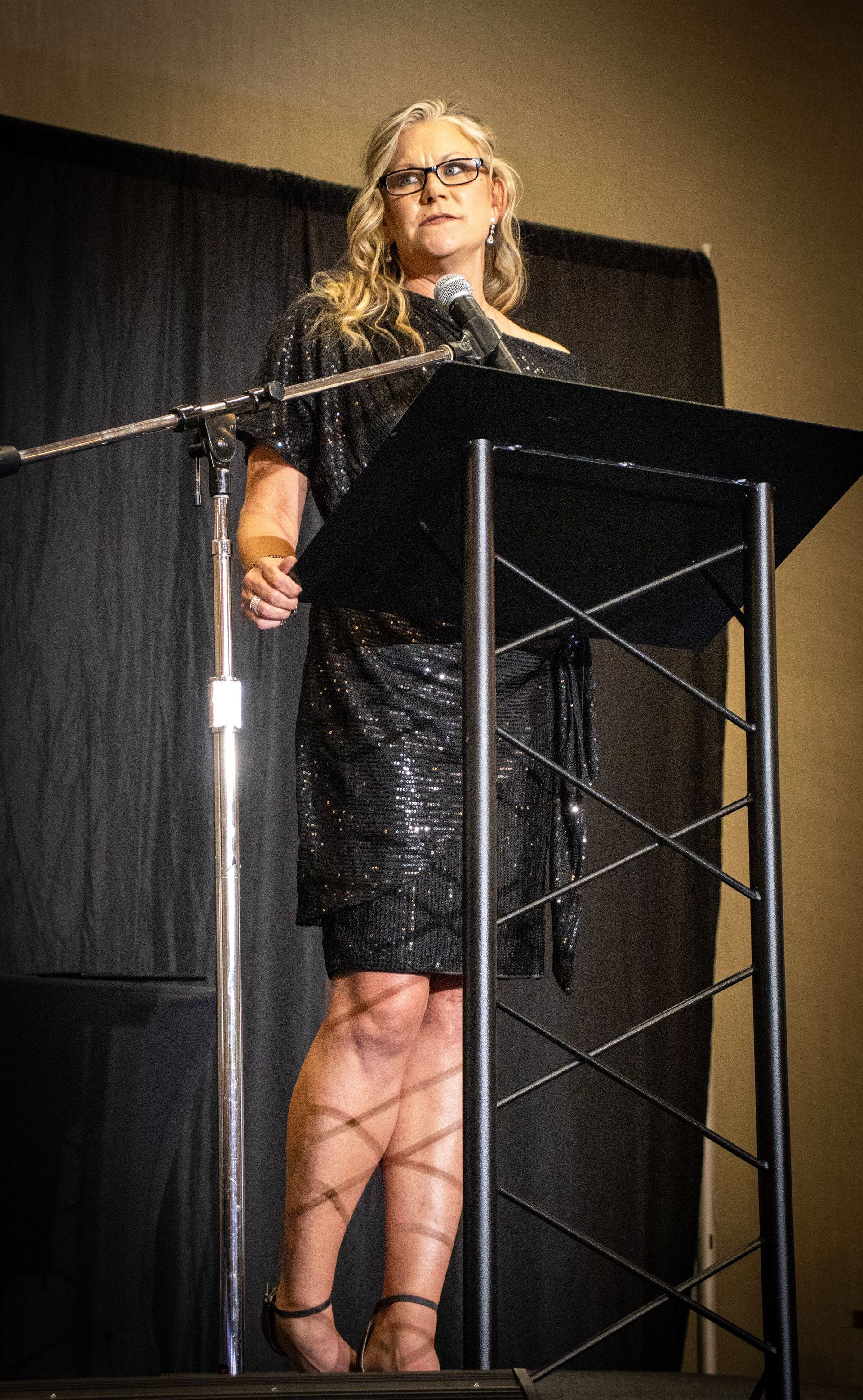 A person with glasses wearing a black dress stands at a lectern, speaking into a microphone against a dark curtain backdrop.