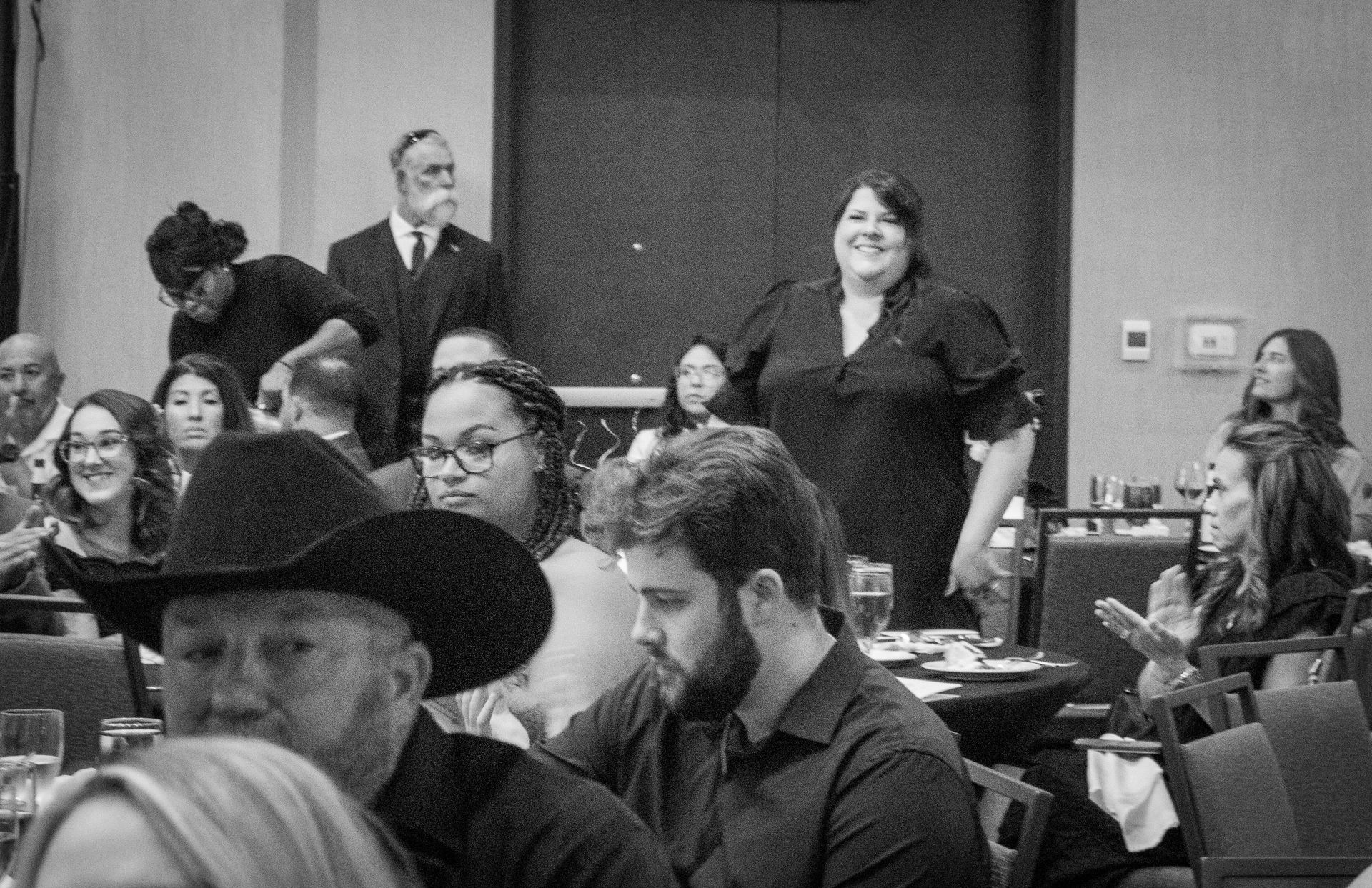 A black-and-white view of a banquet room with guests at tables, including a person in a cowboy hat in the foreground.