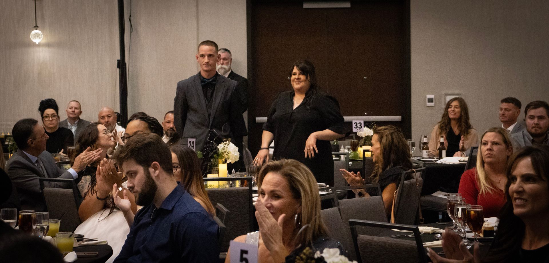 A woman stands at a banquet table while guests seated around her applaud at a dimly lit indoor event.