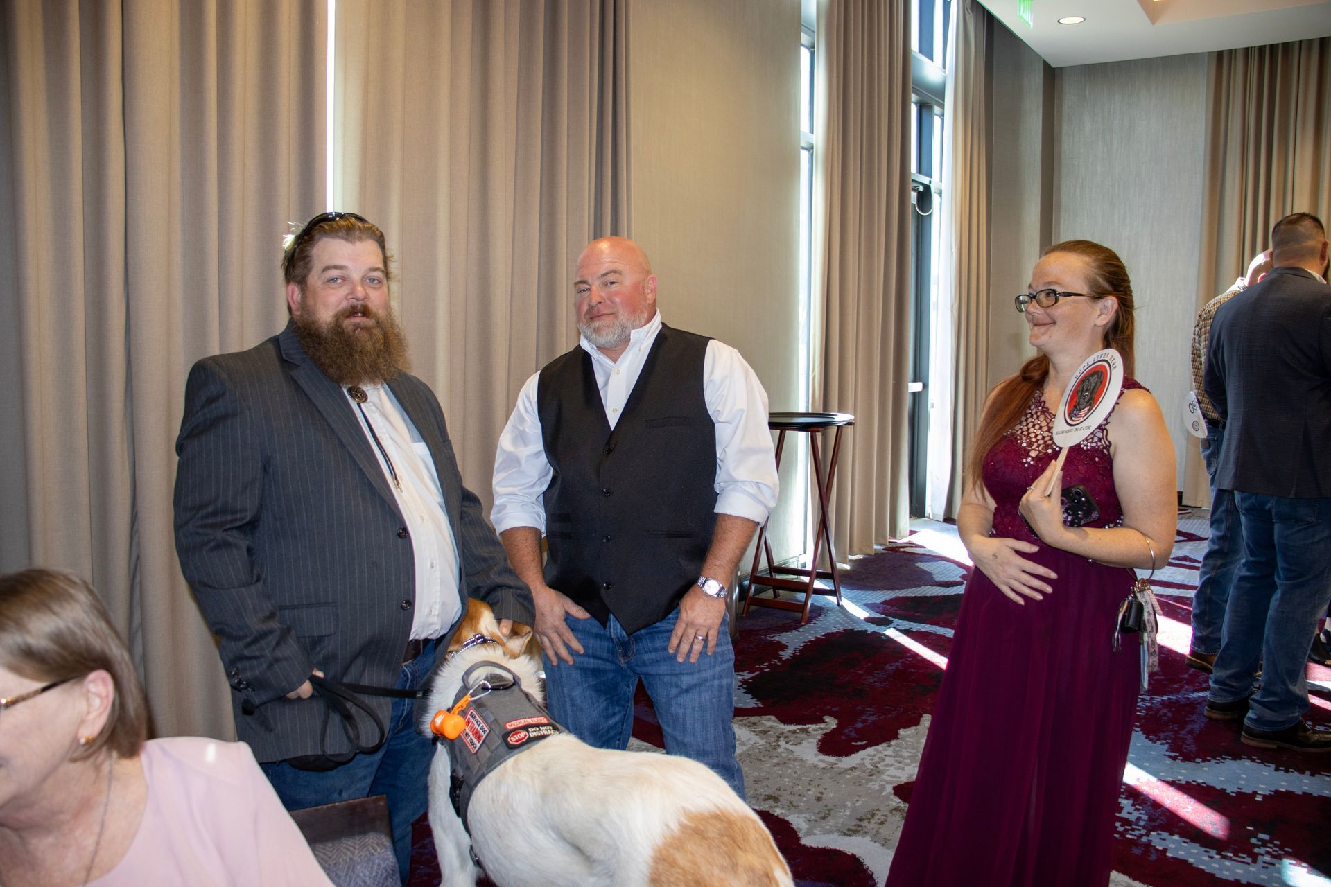 People standing in a room with a dog, including two men in formal attire and a person in a burgundy dress.