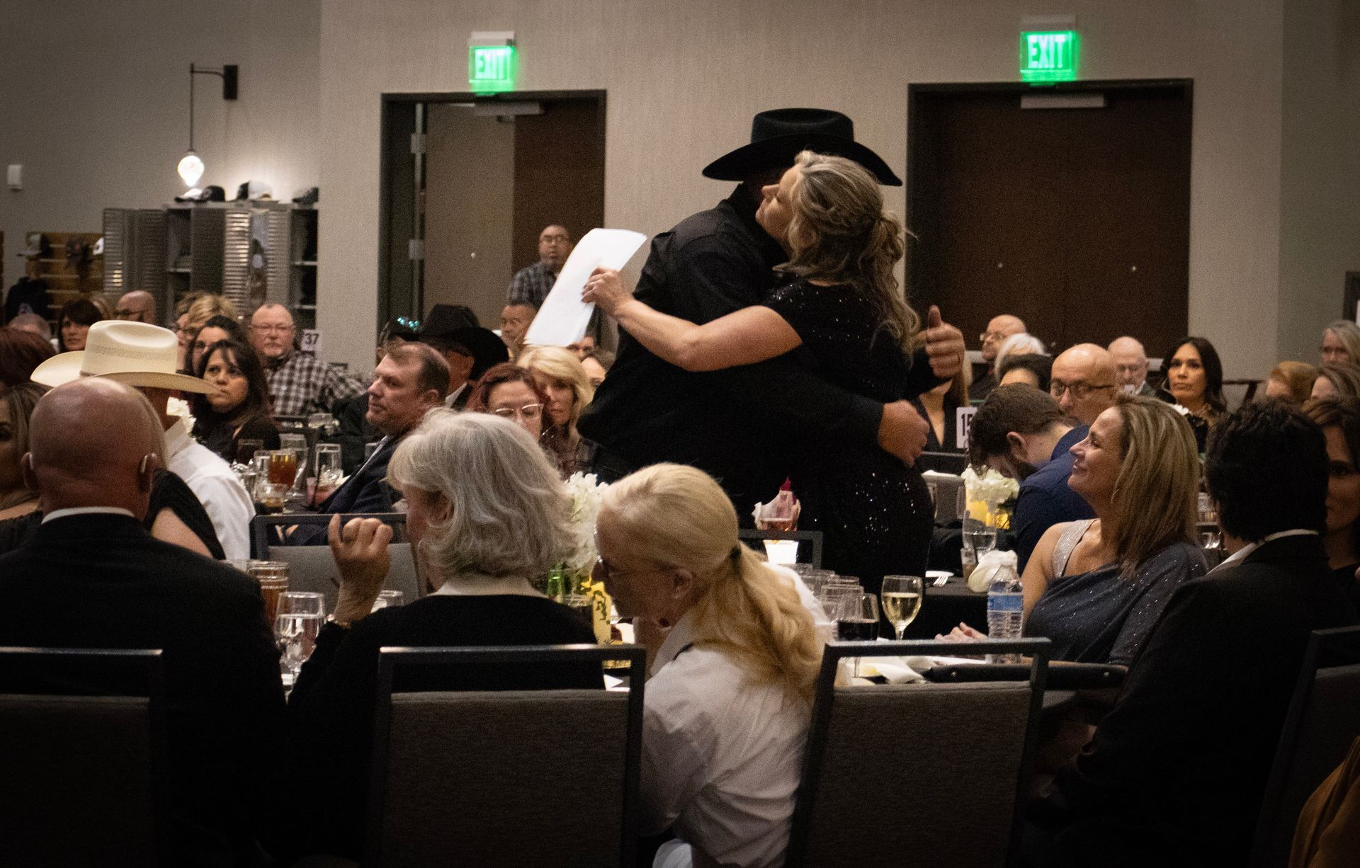 A person in a black cowboy hat hugs another person in a formal ballroom setting filled with guests at tables.
