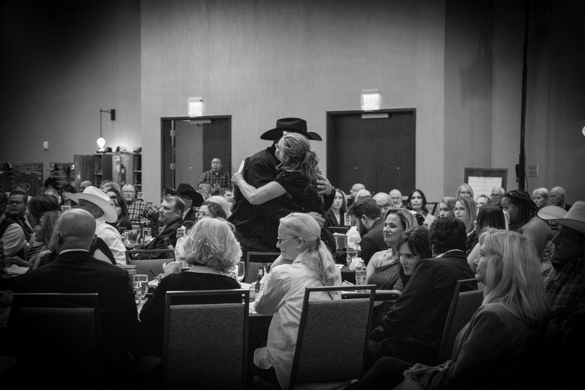 A black-and-white view of a crowded banquet hall where a person in a cowboy hat is embracing someone among the guests.