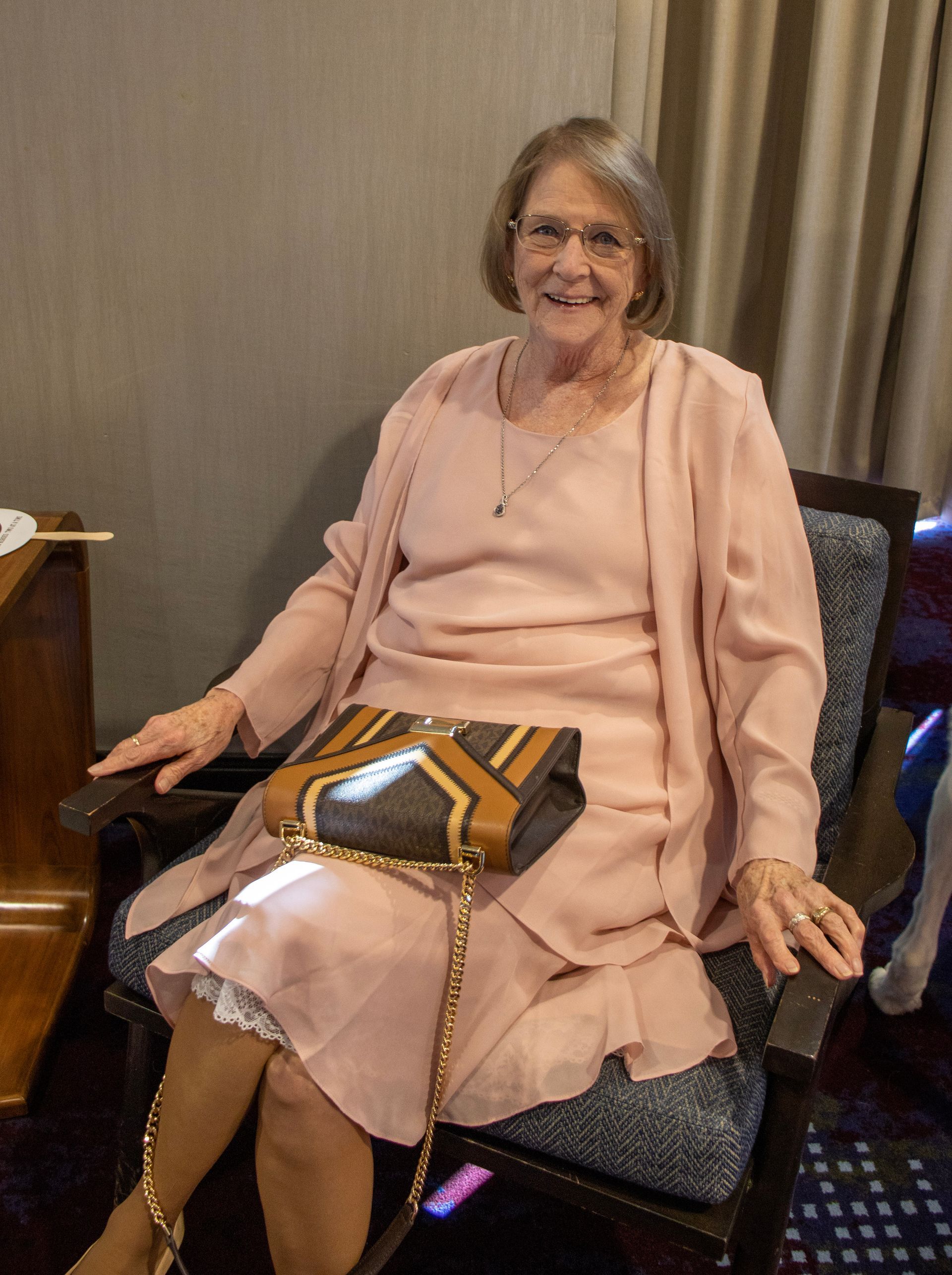 A person in a light pink dress and matching jacket sits in a chair, holding a patterned handbag on their lap.