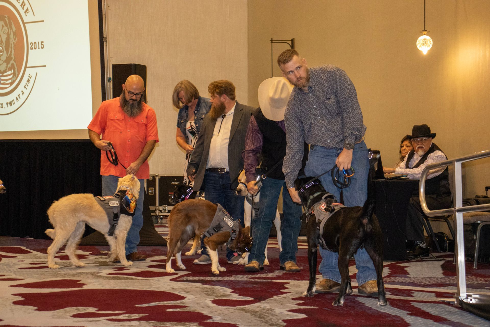 People in a conference room lead three service dogs wearing harnesses across a patterned carpet.