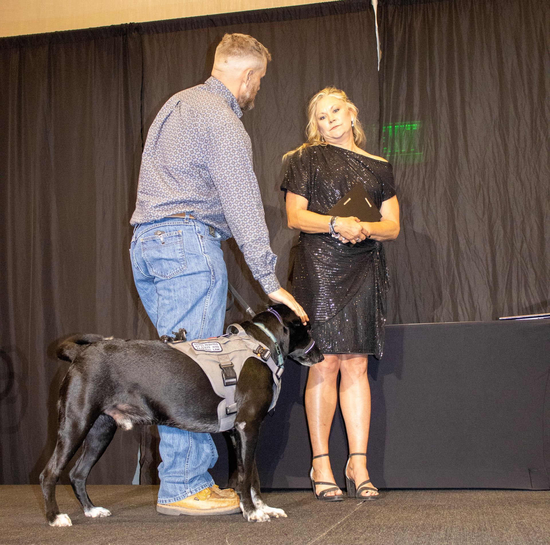 A man in a patterned shirt and jeans pets a black dog in a harness while standing next to a woman in a sequined dress.