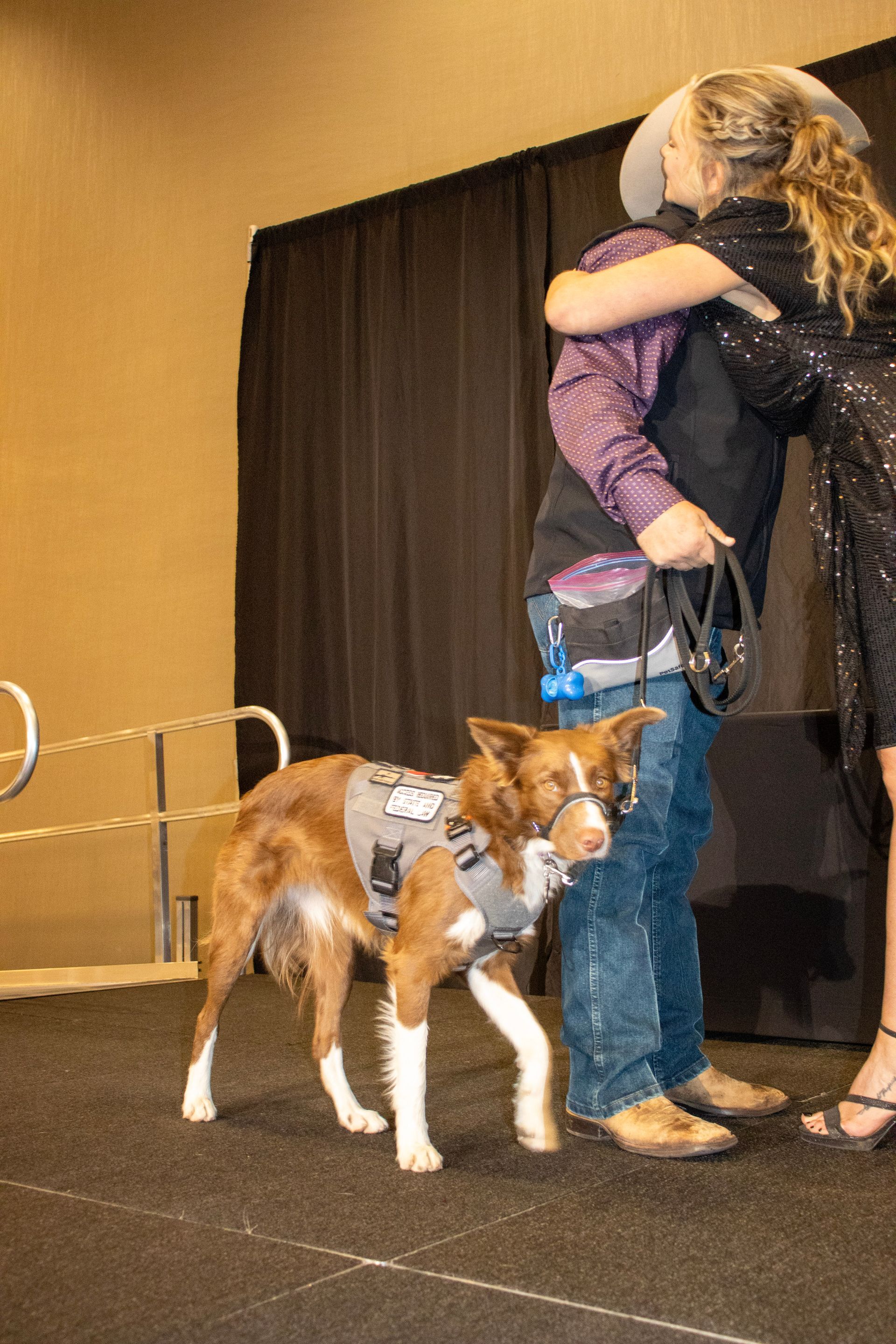A person in a cowboy hat hugs another person while a brown-and-white service dog stands on a stage in front of them.
