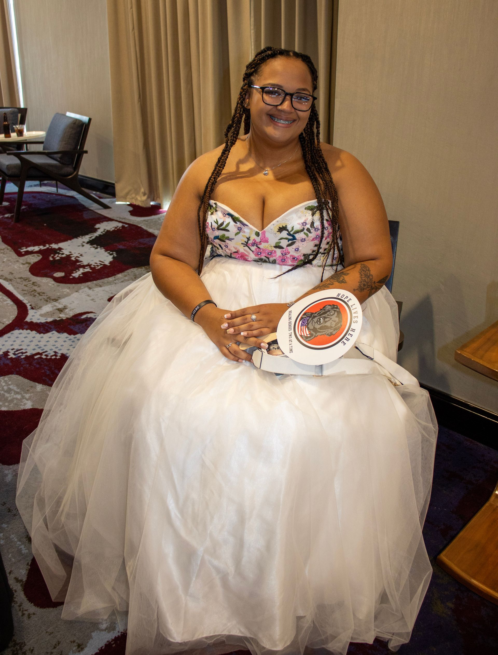 A smiling person wears a floral-print corset and voluminous white skirt, holding a white championship-style belt.