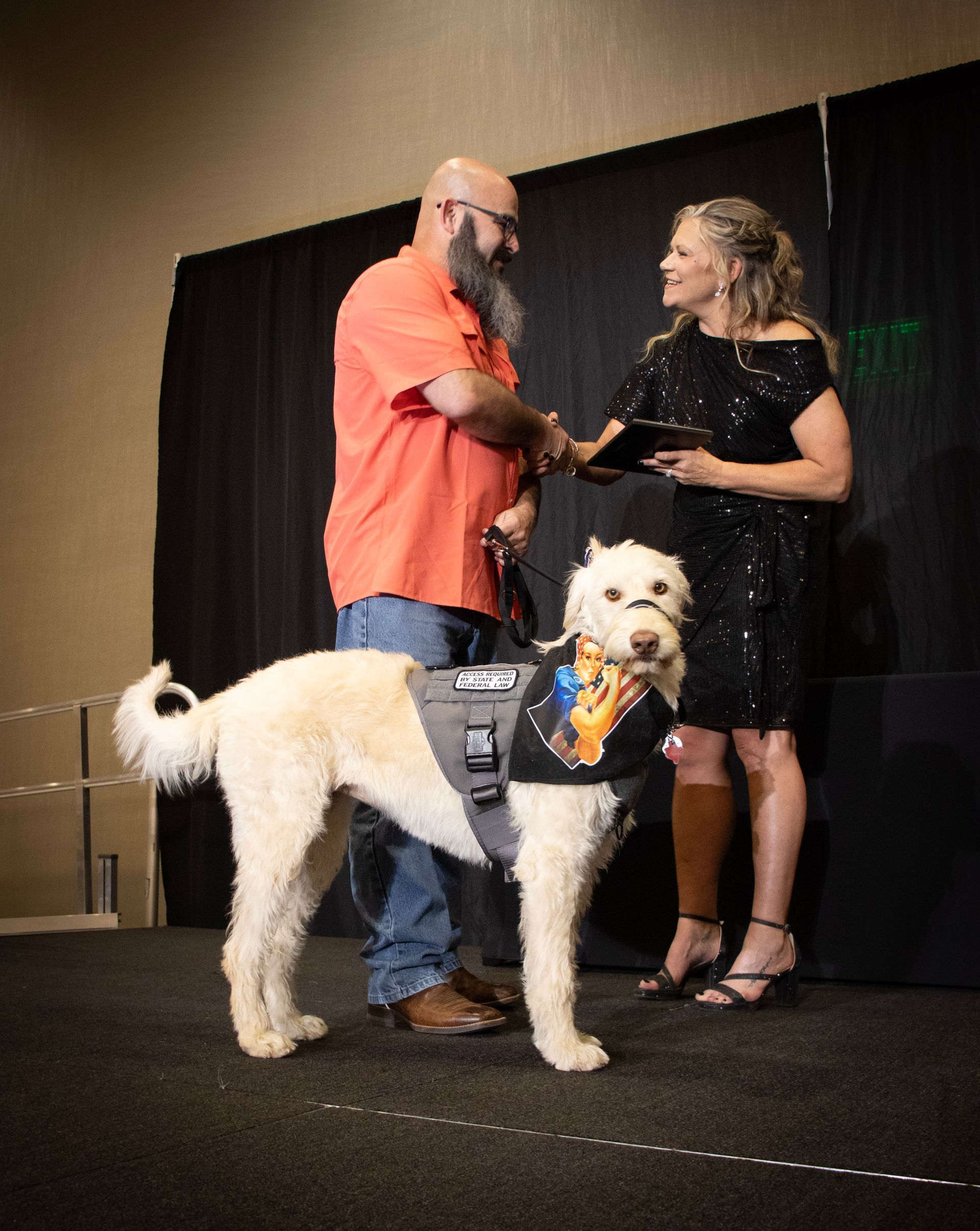 A man in an orange shirt shakes hands with a woman in a black dress on a stage, while a service dog stands between them.