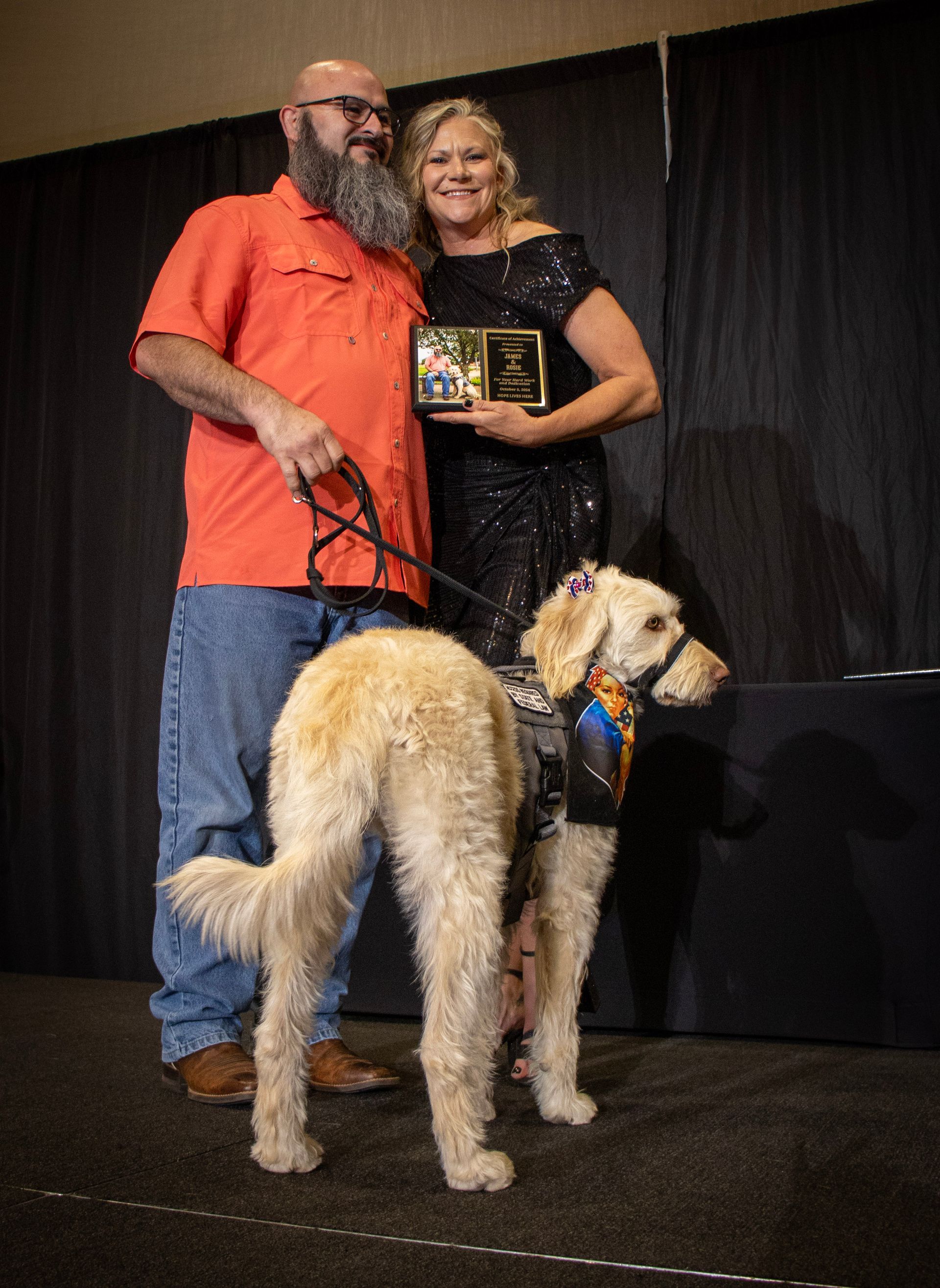 A man and woman holding an award pose with a golden-colored dog wearing a harness in front of a black curtain backdrop.