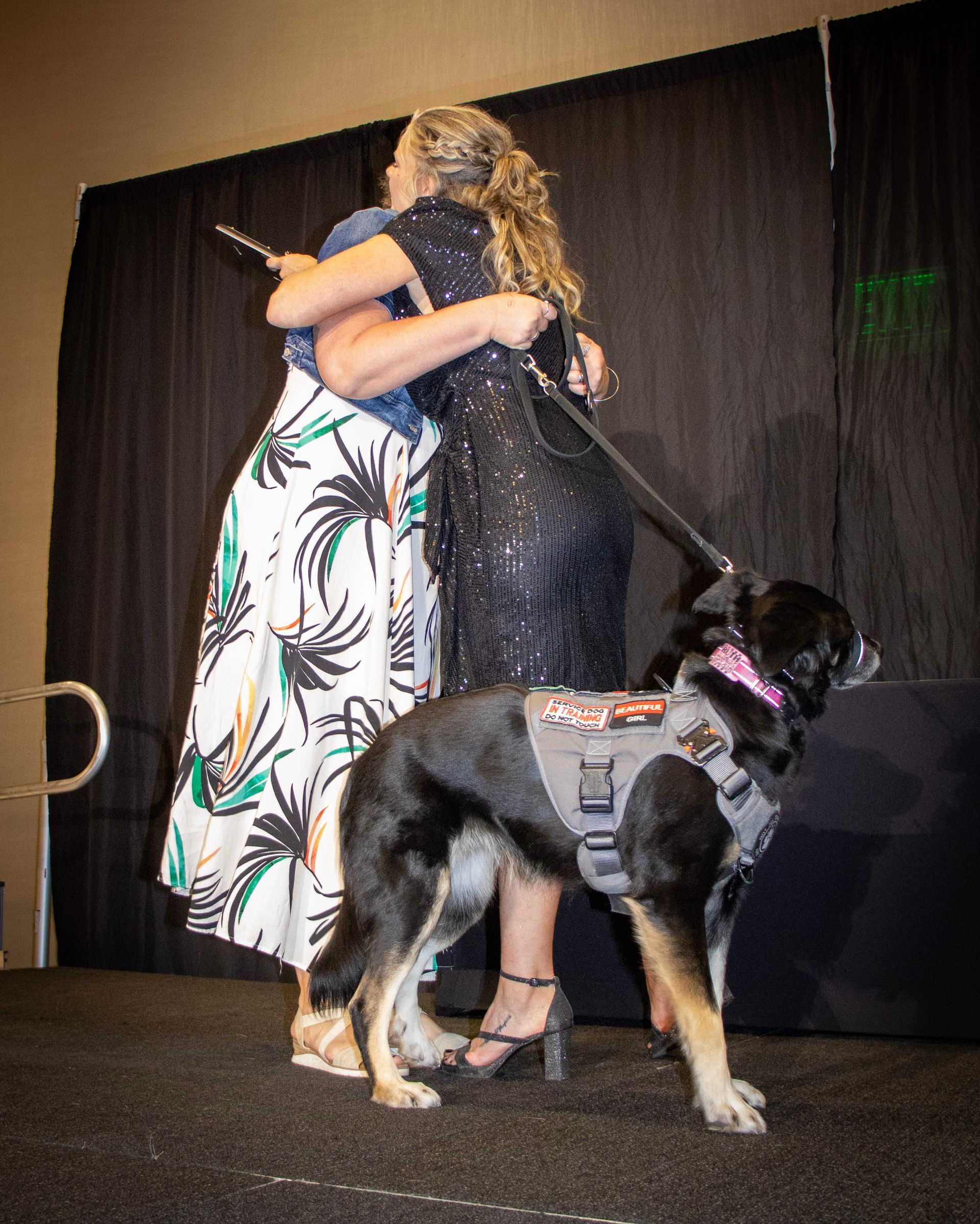 Two people embrace on a stage next to a black and tan dog wearing a grey service harness.