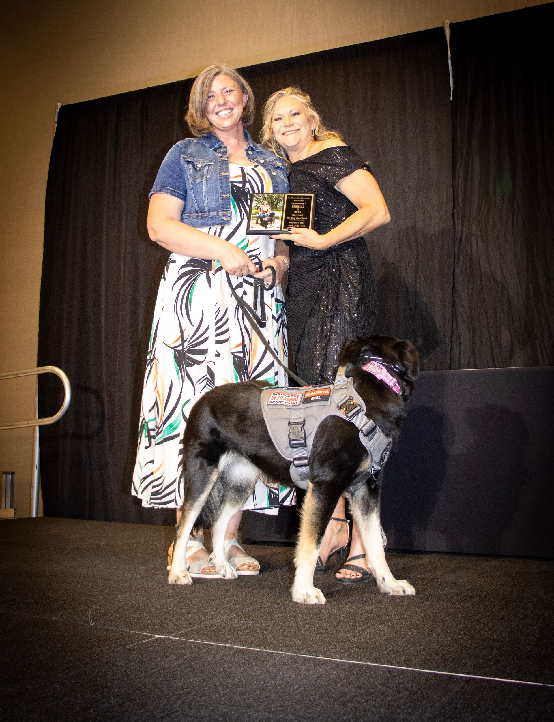 Two people standing on a stage with a service dog wearing a gray vest, holding a small award plaque in front of a curtain.