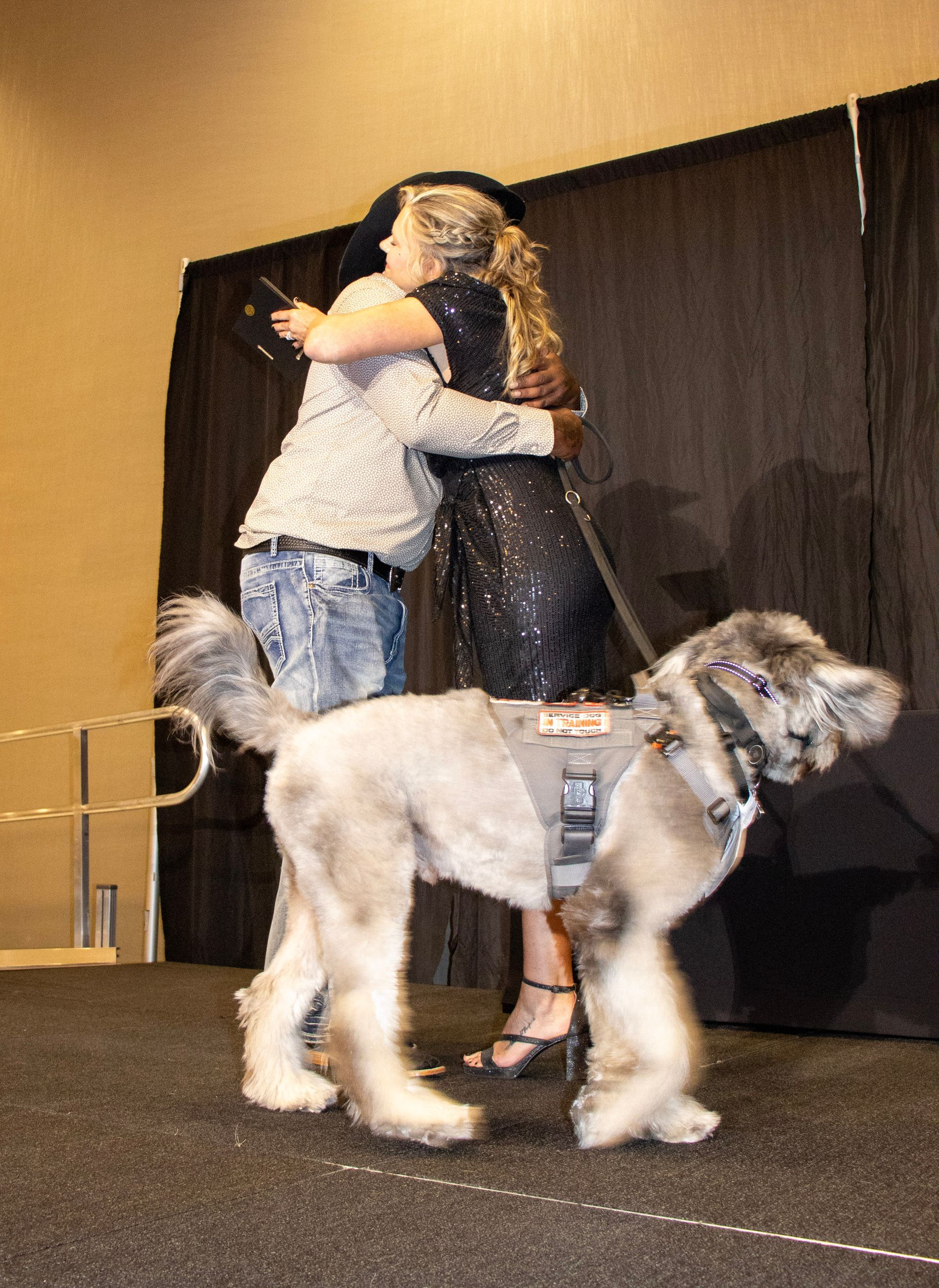 A person in a black dress and a person in a white top hug on a stage, with a grey dog in a harness standing in front.
