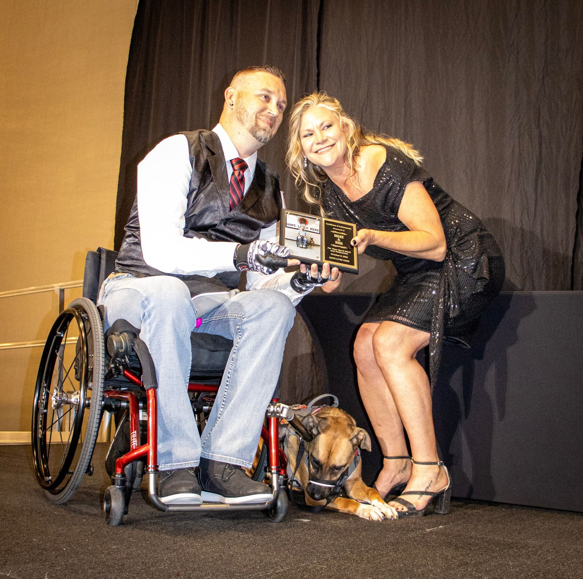 A person in a wheelchair and a person in a black dress holding a plaque, with a dog lying at their feet on a stage.