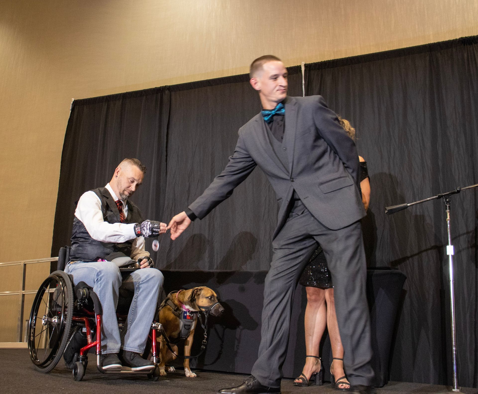 A person in a wheelchair and a person in a suit share a fist bump on a stage with a service dog nearby.
