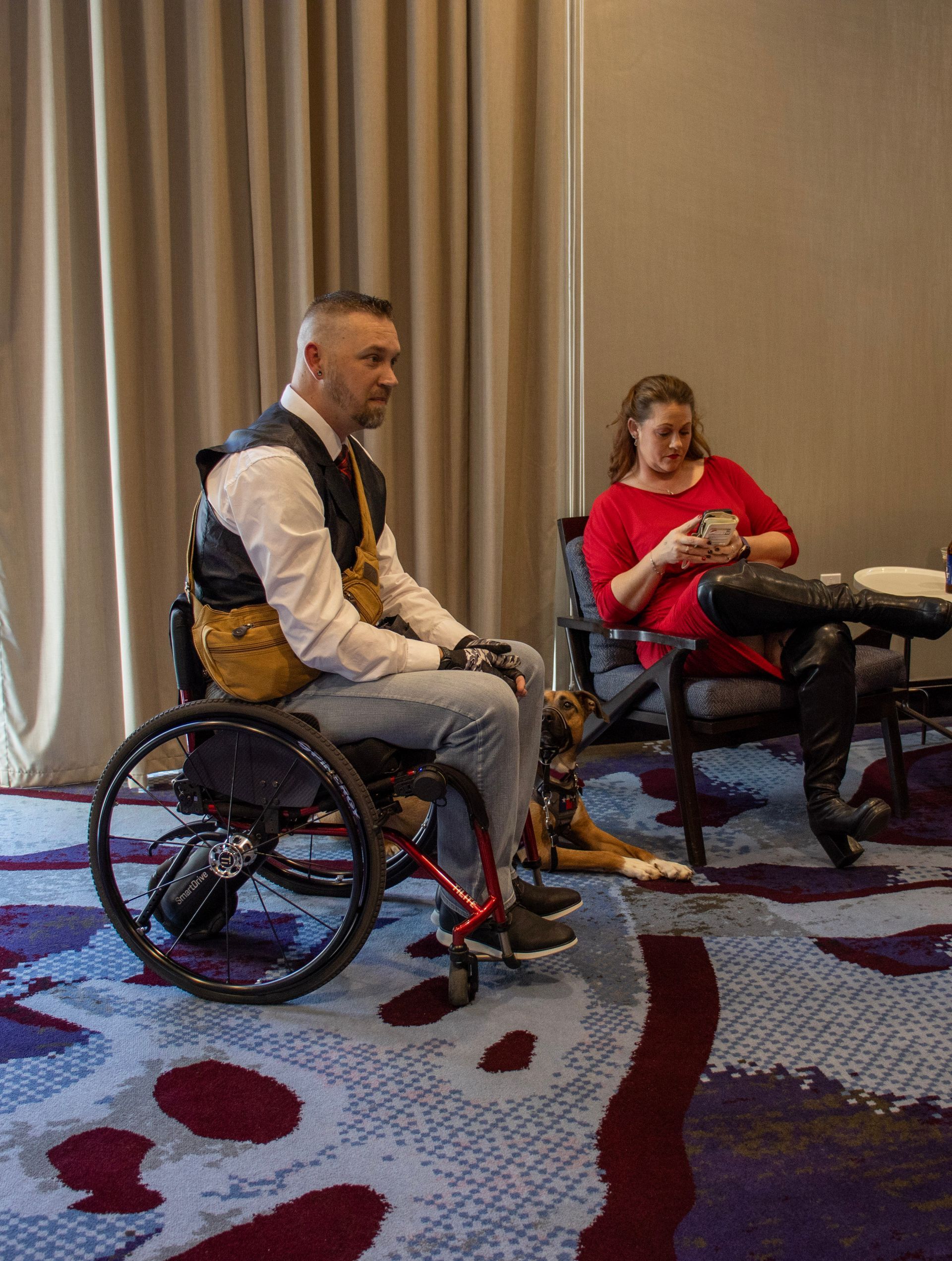 A person in a wheelchair and another sitting nearby look toward the side in a room with patterned carpet and curtains.