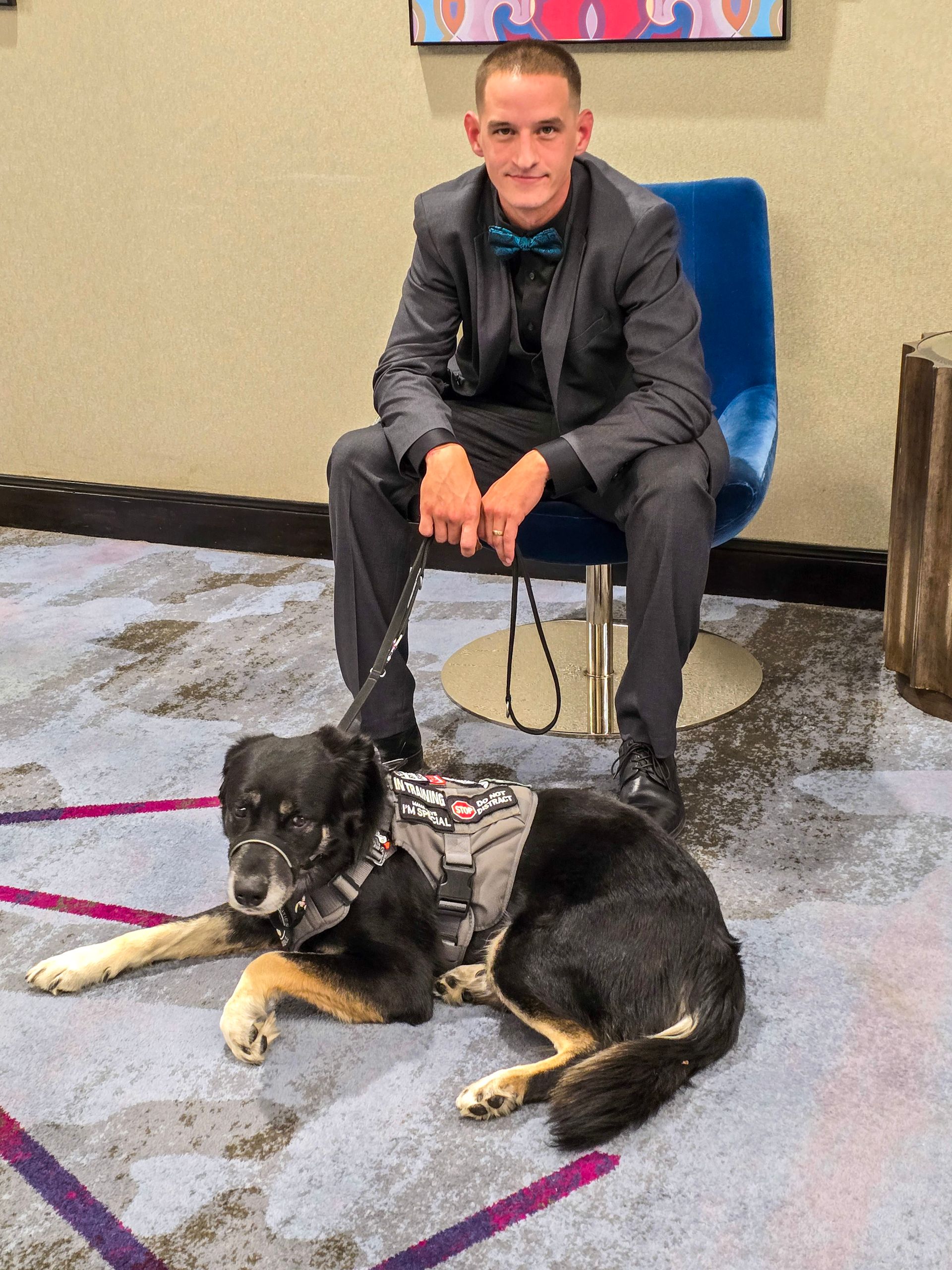 A person in a suit and teal bow tie sits in a blue chair with a service dog wearing a gray harness lying at their feet.