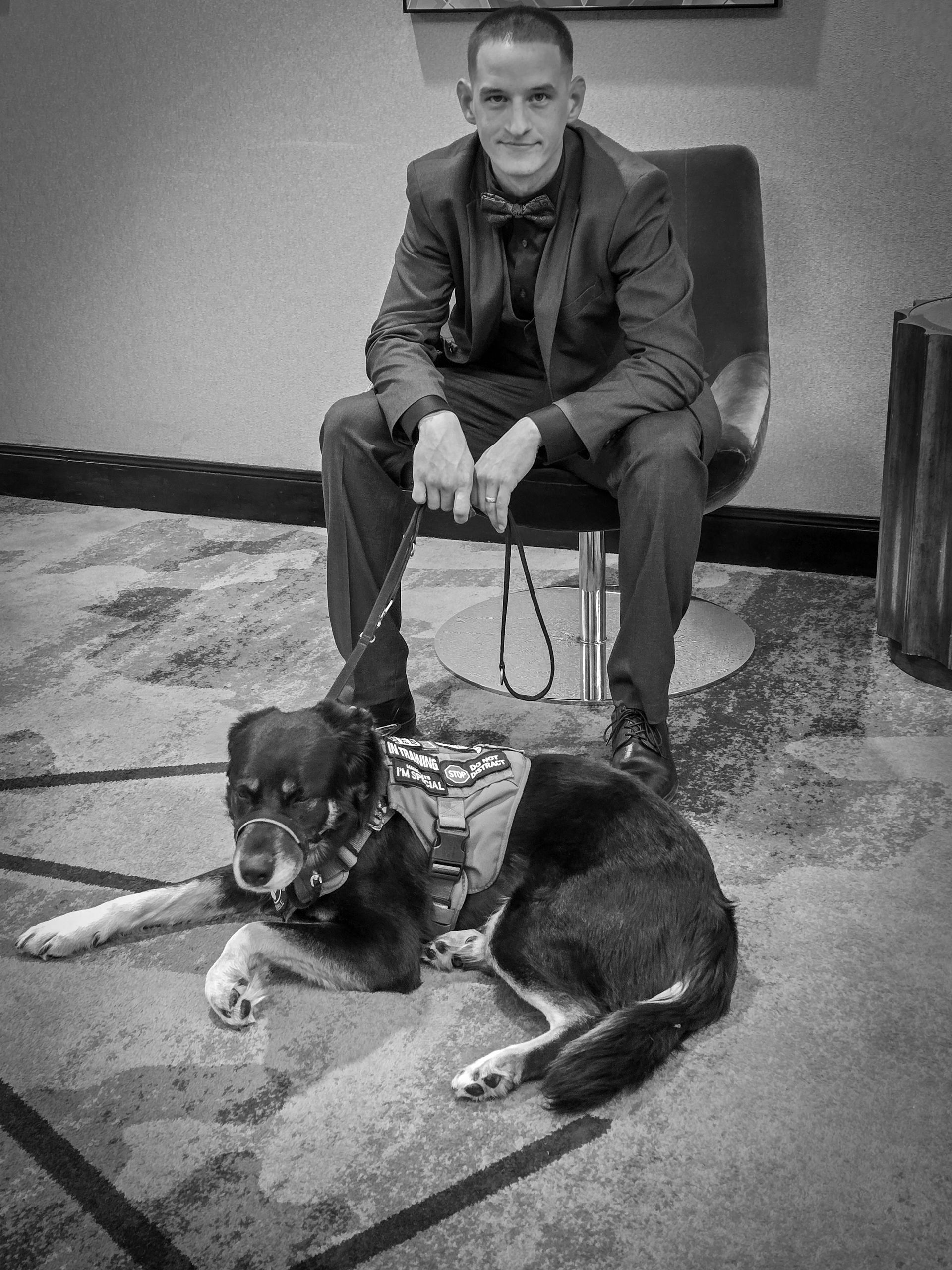 A person in a suit and bow tie sits on a chair, holding the leash of a German shepherd service dog lying on the floor.