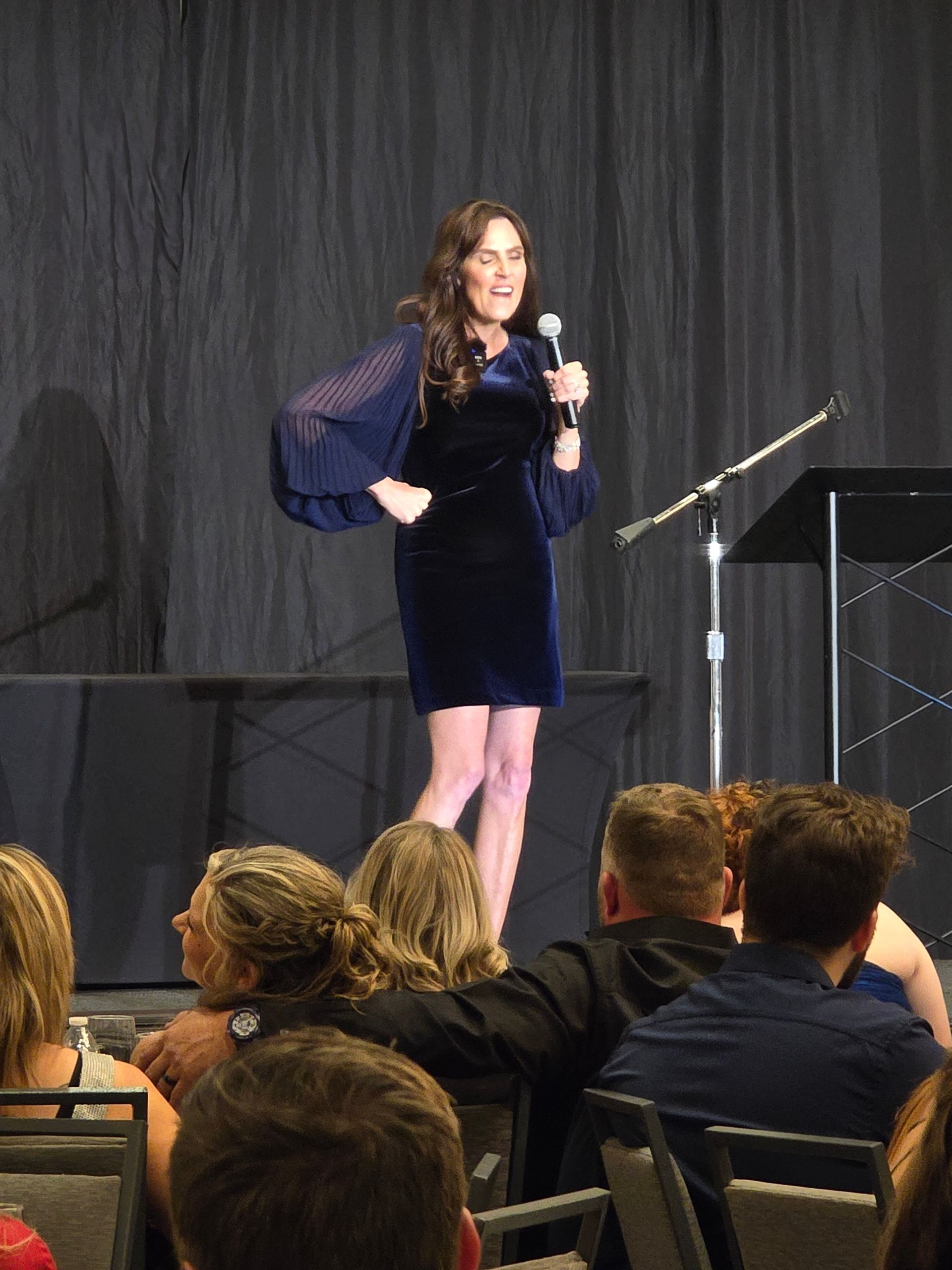 A person in a dark blue, long-sleeved dress speaks into a microphone on a stage with a dark curtain backdrop.