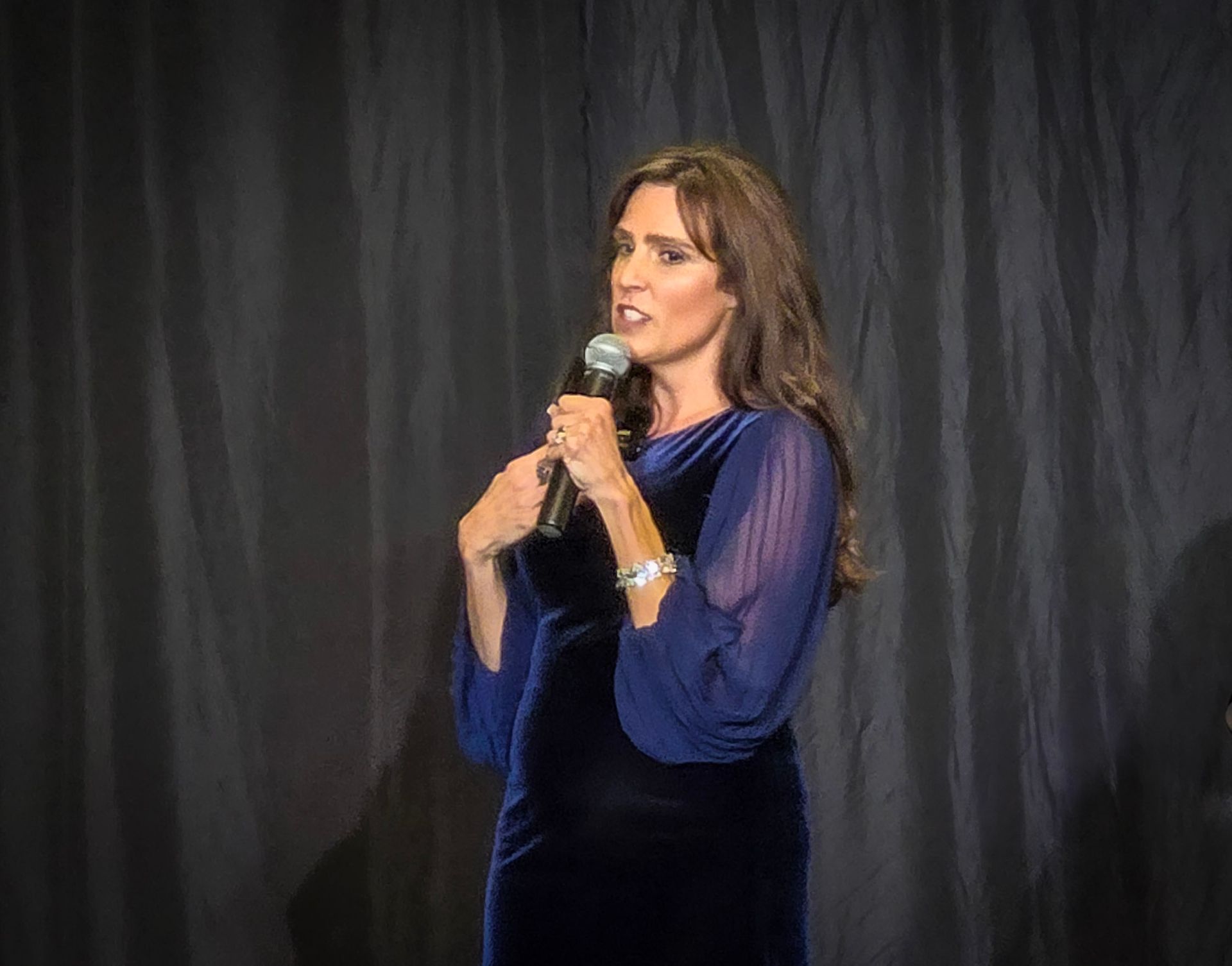 A person speaking into a microphone while wearing a long-sleeved dark blue velvet dress in front of a dark curtain.