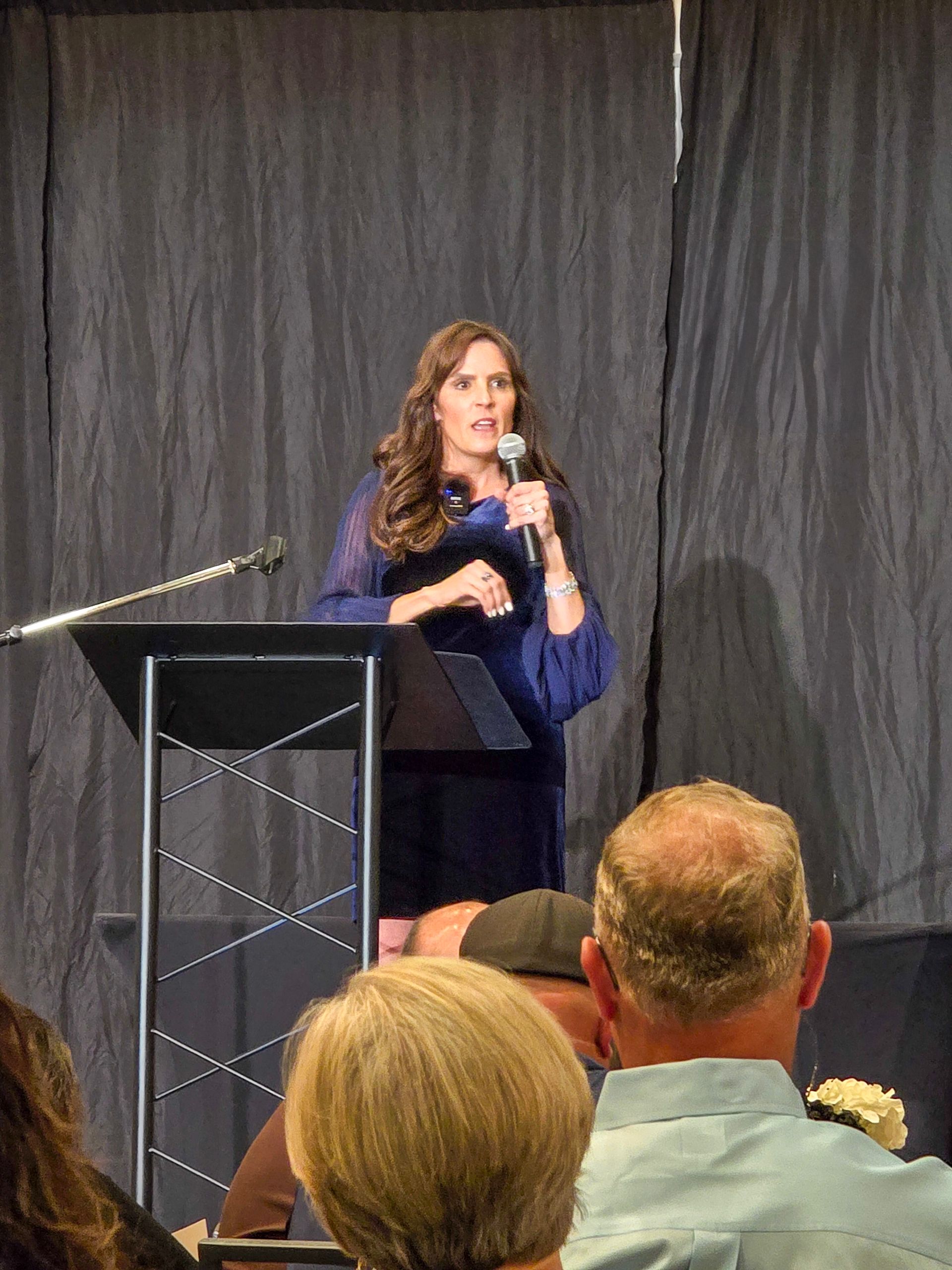 A speaker in a dark blue blouse stands at a podium behind a microphone, addressing an audience in a dimly lit room.