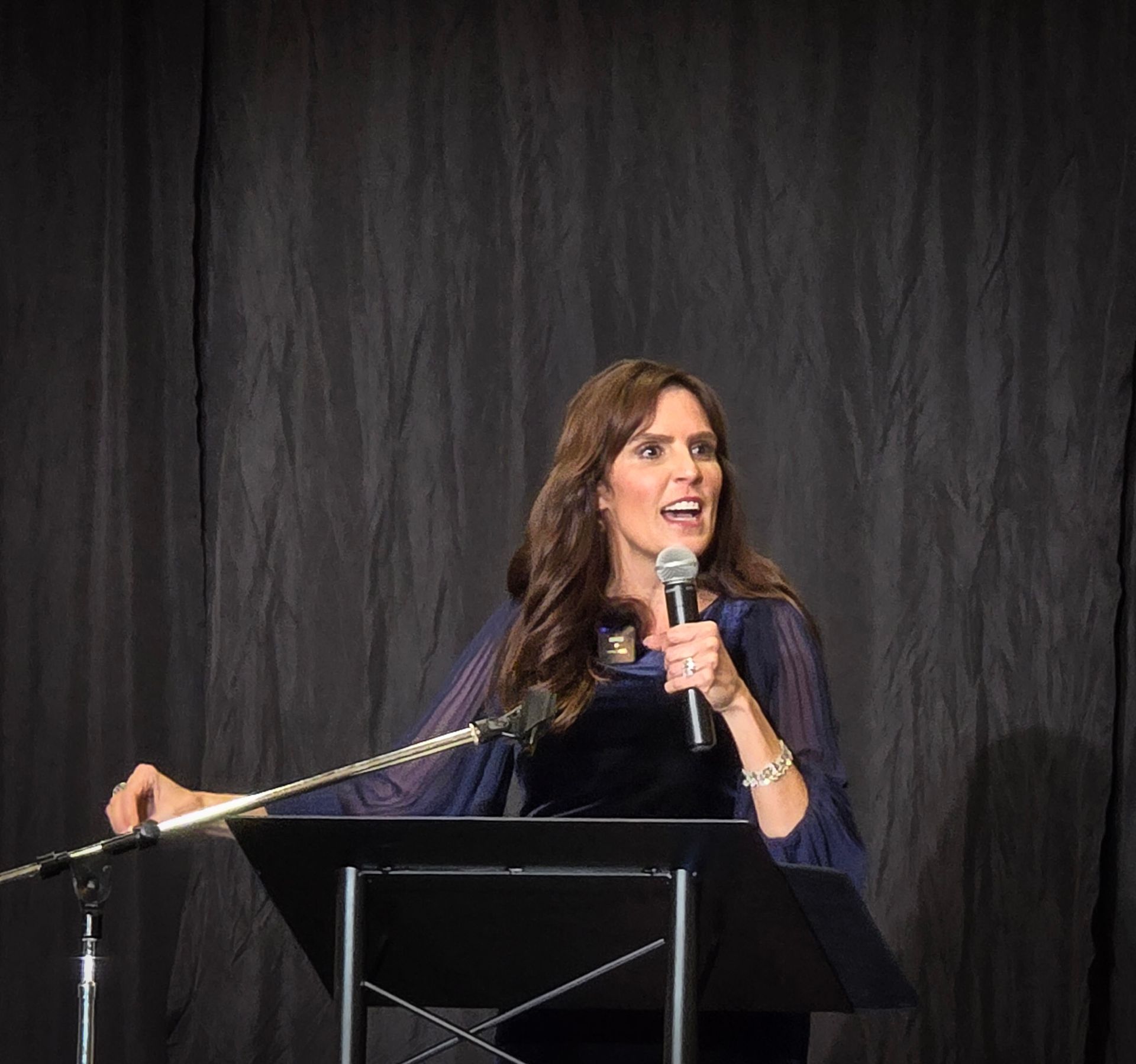 A woman speaking into a microphone while standing at a podium in front of a dark, wrinkled fabric backdrop.