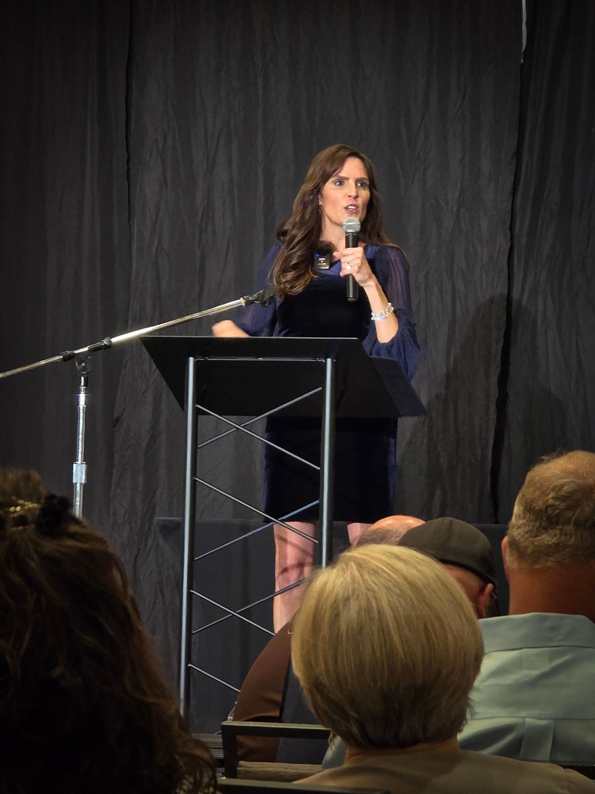 A woman speaks into a microphone while standing at a podium in front of a dark curtain, viewed from an audience.
