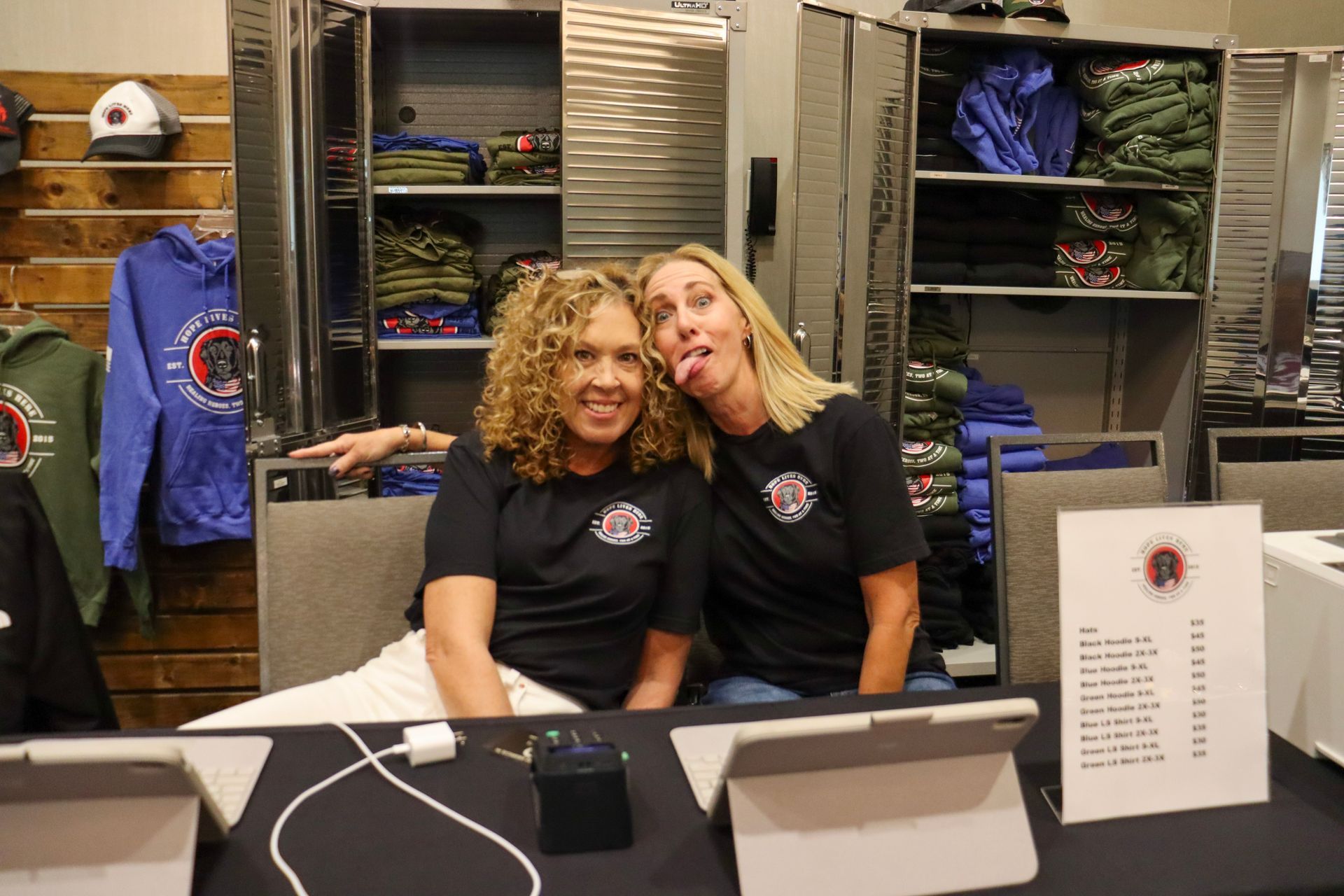 Two people in matching black t-shirts sit at a merchandise table in front of storage lockers; one sticks their tongue out.