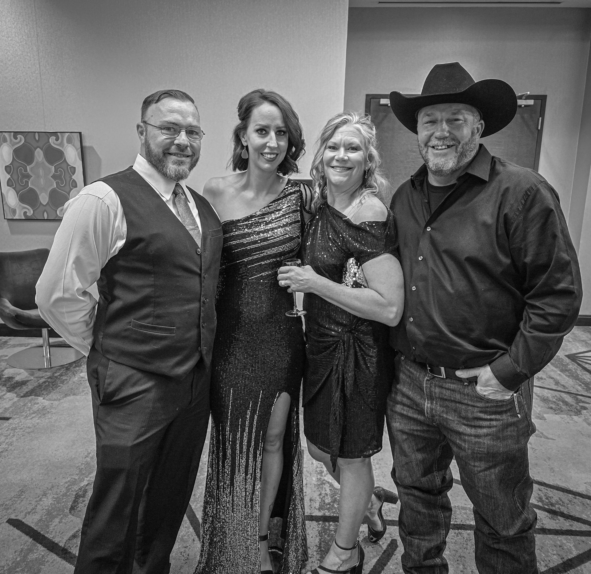 Four adults stand smiling side-by-side in a room, dressed in formal attire including suits, a gown, and a cowboy hat.